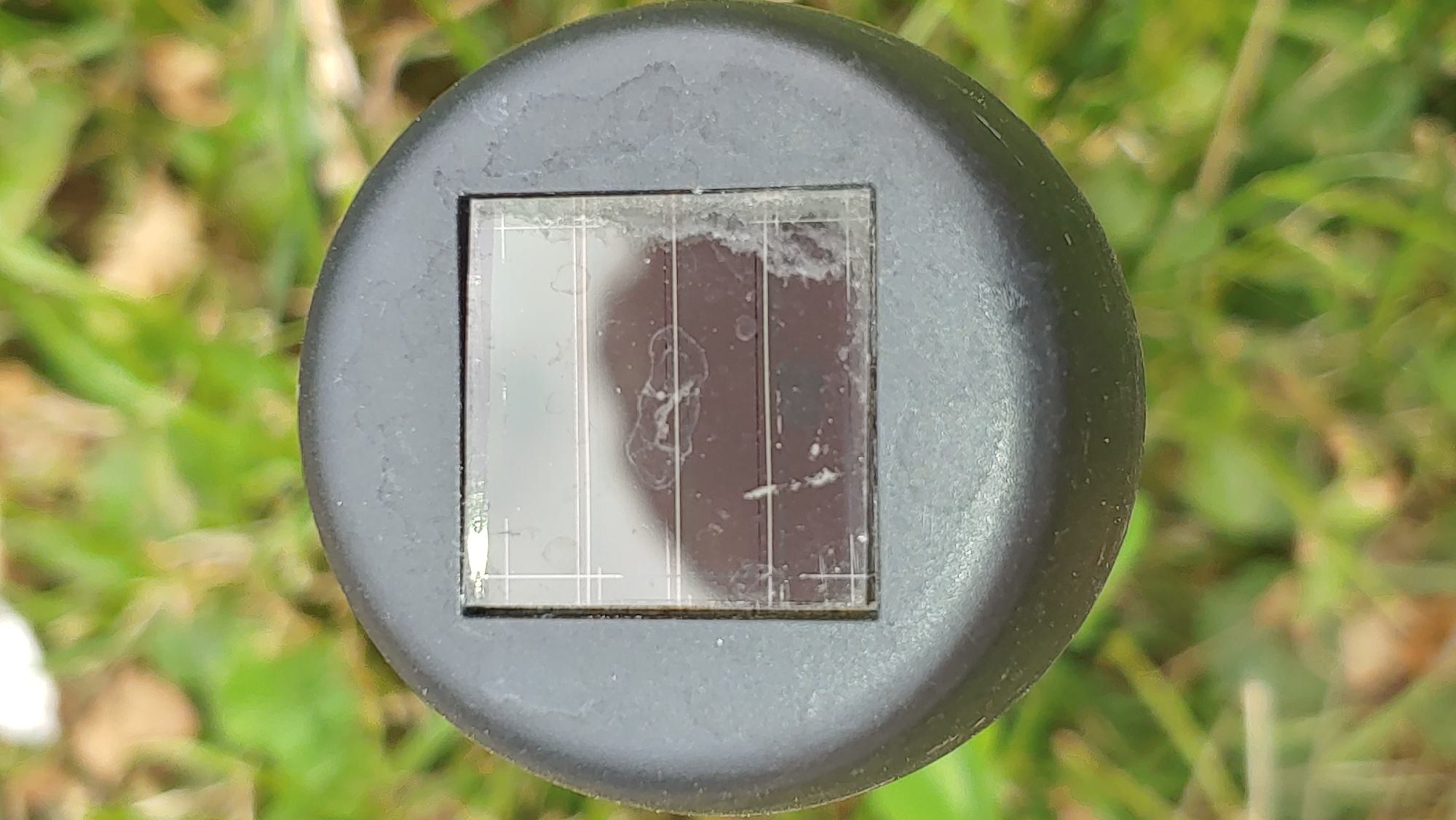 The image shows a close-up, top-down view of a small, square solar panel set into a black, circular casing. The solar panel is slightly dirty and appears to have some smudges or water marks on its surface. The casing is part of a larger object, likely a garden light or similar outdoor fixture, that is positioned on a background of out-of-focus green grass and some brown leaves. There are no people or discernible actions occurring in the image. The location is not identifiable from the visual information provided.