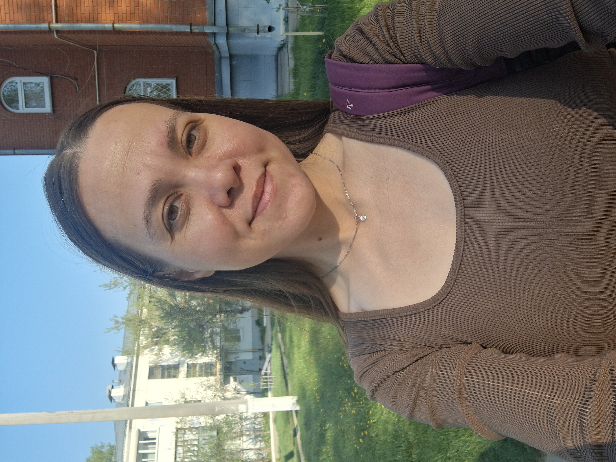 A woman with long brown hair is pictured in a selfie, wearing a brown shirt and a purple strap of a bag. The background shows a red brick building with arched windows and a white building with multiple windows. The location is likely the city of Chaikovsky, Russia. Green grass and trees are also visible in the scene.