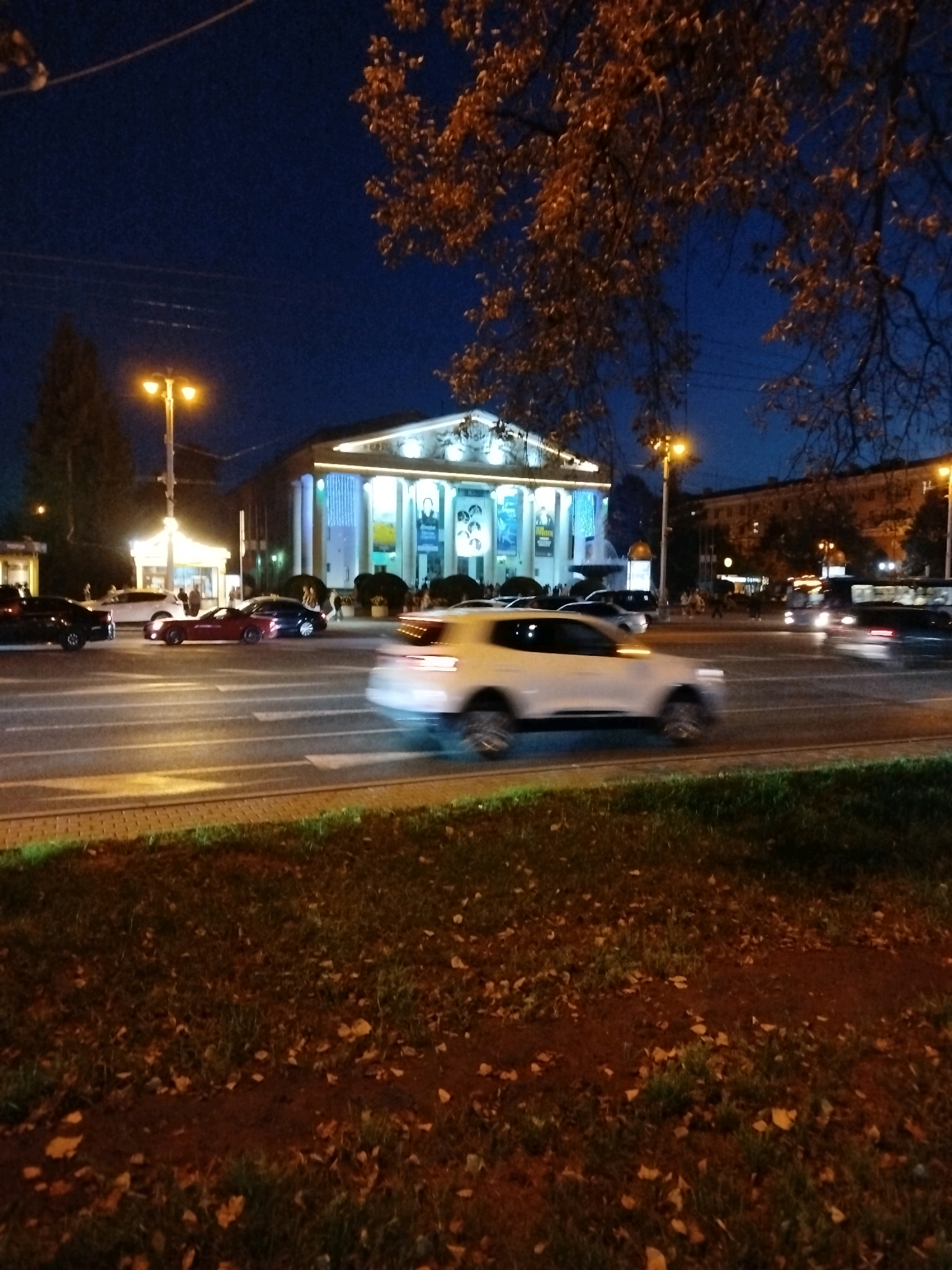 The image depicts a nighttime scene on a city street in Kemerovo, Russia, likely in front of a cultural institution such as a theater or palace of culture. A large building with illuminated columns and signage is visible in the background, suggesting it is an important public structure. The street is active with several cars, some of which are blurred due to motion, indicating traffic. Streetlights cast a warm glow on the scene, and autumn foliage from trees in the foreground and middle ground adds a seasonal element. The ground is covered with grass and fallen leaves, typical of an urban park or roadside area.