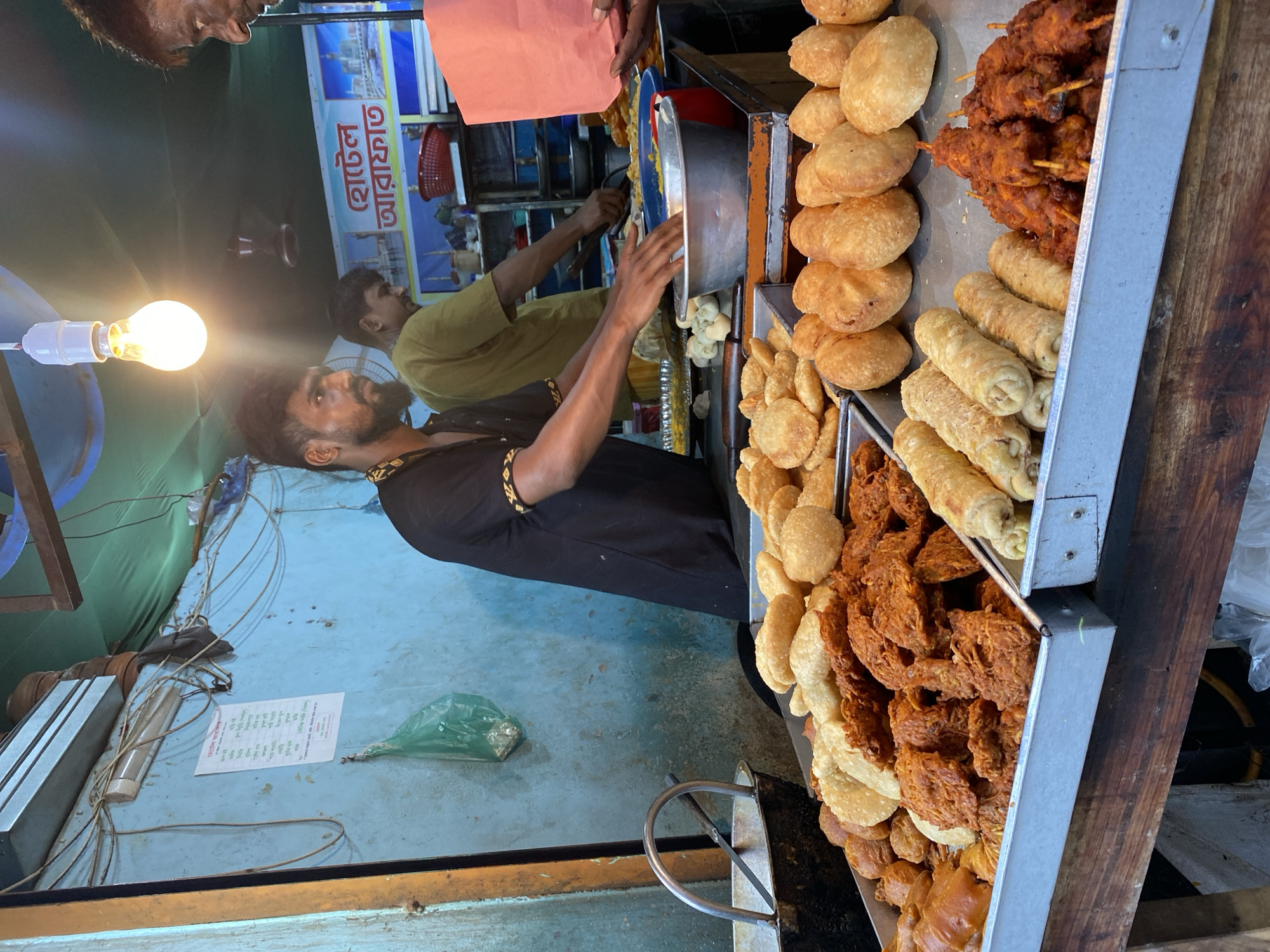 The image depicts an outdoor food stall in Dhaka, Bangladesh, likely a street food vendor. The stall is set up on a wooden counter with several trays of fried food items. In the foreground, a tray is filled with an assortment of fried snacks, including what appear to be samosas, pakoras, and possibly puri. To the right, another tray contains spring rolls and skewers of red-colored fried chicken or meat. In the background, a man with a beard wearing a black shirt with a patterned collar is attending to the food, his hands near a metal bowl. Another man in a yellow-green shirt is visible behind him, also working. A bright light bulb hangs overhead, illuminating the scene. The stall has a blue-painted wall behind it, with some wiring and a printed menu or advertisement. A red basket is visible on a shelf. The overall impression is of a busy, casual food service environment characteristic of street food culture in Dhaka.
