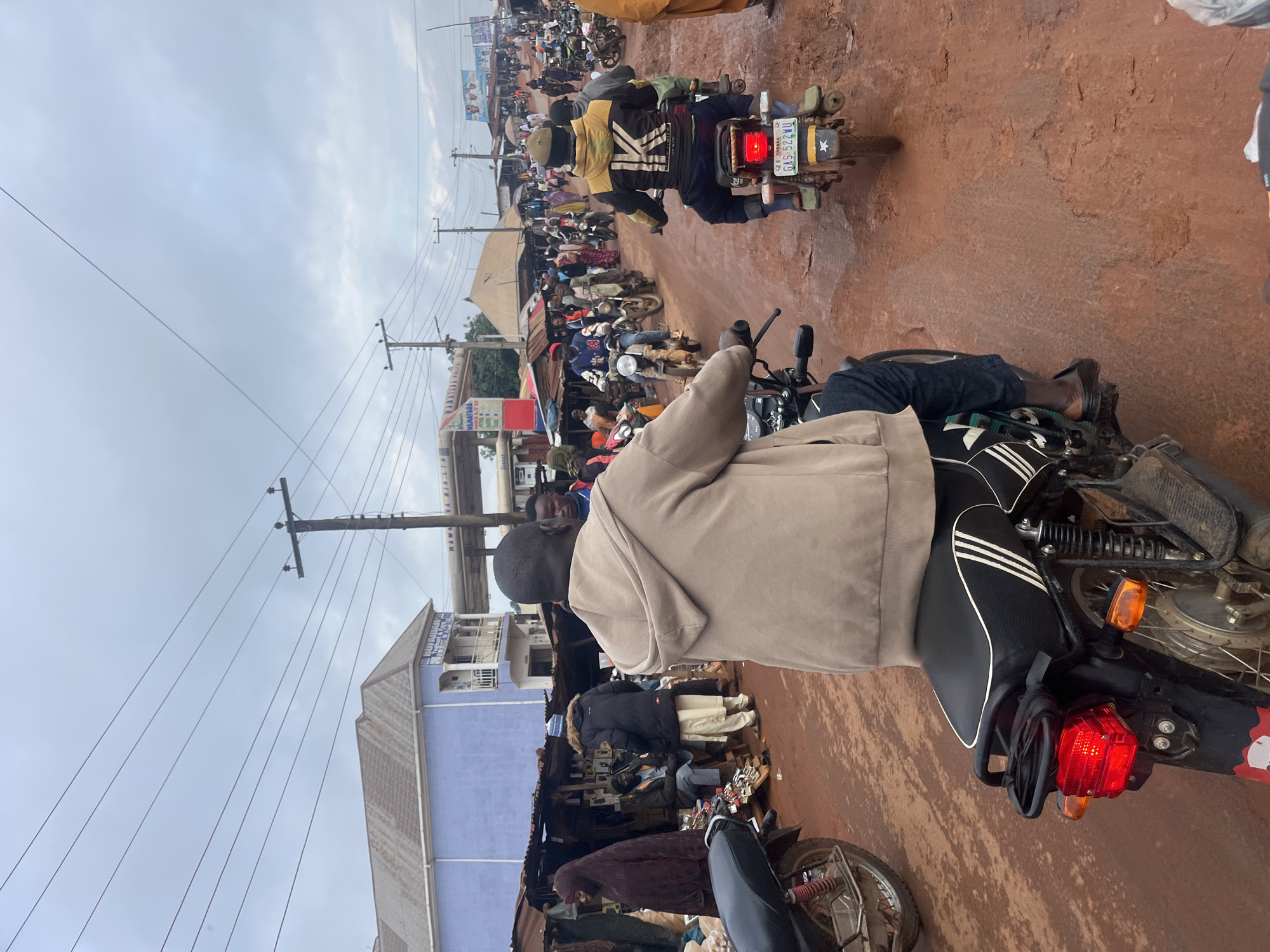 The image depicts a busy street scene in Gembu, Nigeria, likely during the day given the bright, overcast sky. The primary activity is a congregation of motorcycles and people, suggesting heavy traffic or a market day.
The foreground features the back of a person on a motorcycle, with their legs and feet visible. The motorcycle itself is dark with white stripes on the seat and a prominent red taillight. The ground is a reddish-brown, indicative of dirt or unpaved roads common in the region.
In the mid-ground and background, numerous other motorcycles are parked or in motion, with many people, some wearing traditional clothing, gathered around. There are stalls and shops lining the street, suggesting a commercial area. One building has a blue facade with white architectural details. Overhead, utility poles and wires are visible, characteristic of a developed area.
There is some visible text on a license plate of a motorcycle in the mid-ground, reading "GAS 5224 U" and a star emblem. Other signs are too blurry to read clearly, but their presence indicates ongoing commerce. The overall atmosphere appears lively and active, with a sense of community engagement in a marketplace setting. The overcast sky suggests potentially humid or rainy weather.