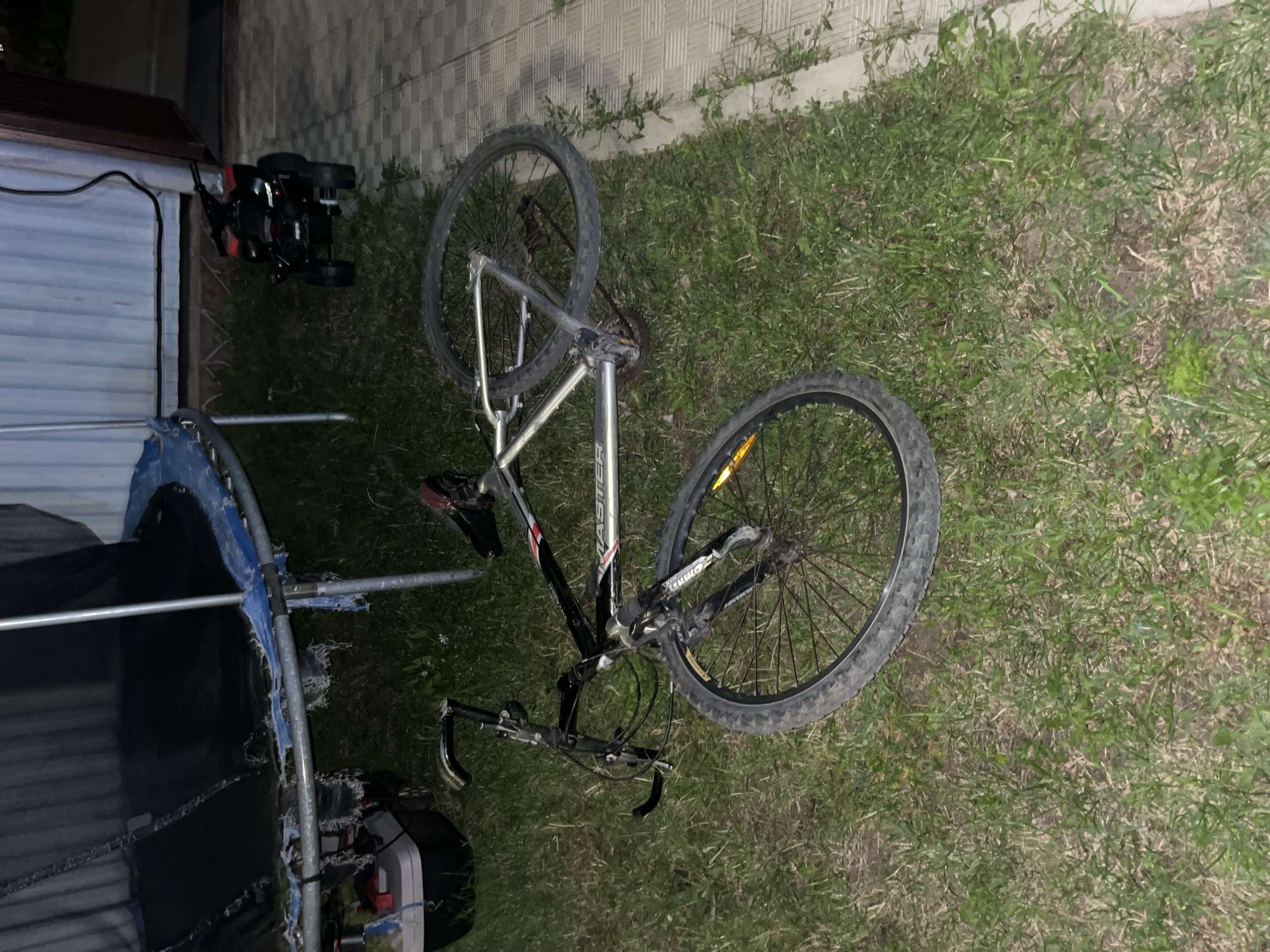 A bicycle lies on its side on a patch of grass next to a paved walkway. The bicycle is silver and black, with a black seat and handlebars. It appears to be a mountain bike. Behind the bicycle, a trampoline with a torn mat is partially visible. To the right of the trampoline and behind the bicycle, a small, black, electric ATV is parked on the paved walkway. The scene appears to be outdoors, possibly in a backyard or park. There are no people visible in the image. The image quality is somewhat grainy, suggesting it was taken at night or in low light conditions. There is no information about the location being Saratov, Russia.