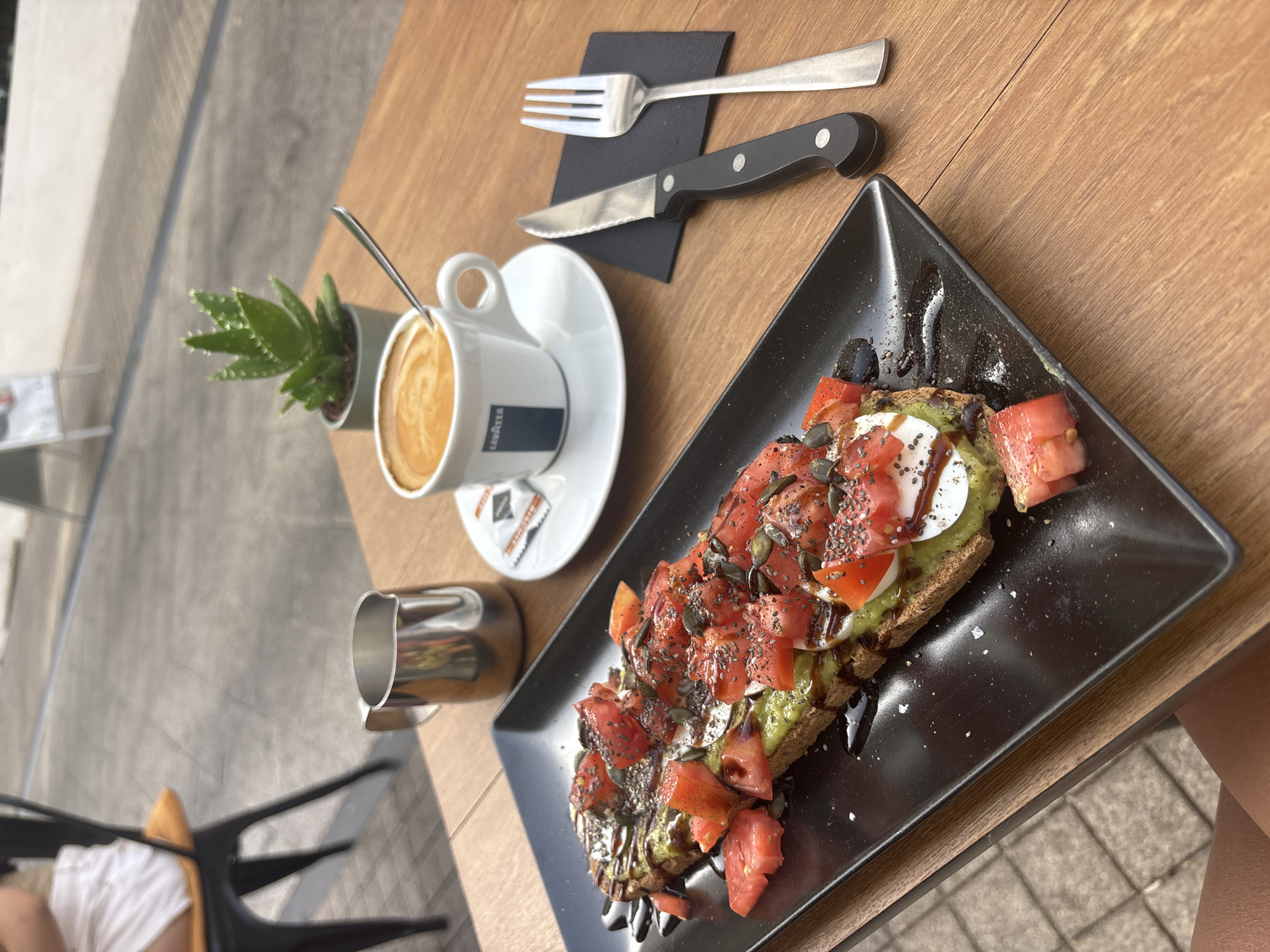 An overhead, medium shot captures a table setting outdoors in Marbella, Spain, featuring a close-up view of a savory avocado toast and a cappuccino. The avocado toast, presented on a rectangular black plate, is generously topped with chopped tomatoes, mozzarella, and sprinkled with chia seeds and pumpkin seeds, all drizzled with balsamic glaze. Beside the toast, a white ceramic cup and saucer hold a cappuccino, with a small silver spoon resting on the saucer. A miniature stainless steel pitcher is positioned behind the toast. To the right of the cup, a black-handled knife and a silver fork rest on a small black napkin. In the background, a small potted succulent plant sits near the coffee cup, and the blurred outlines of a chair and outdoor seating area suggest a café or restaurant environment. The wooden table surface and paved ground indicate an outdoor seating area.