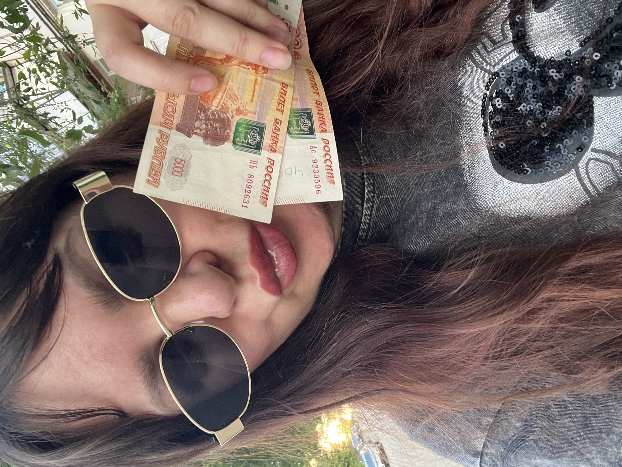 The image depicts a close-up, slightly upward-angled outdoor selfie of an individual, stated to be located in the city of Chaikovsky, Russia. The person, likely female, has long, dark reddish-brown hair, parted on the side, falling over her shoulders and down her chest. She wears dark, oval-shaped sunglasses with gold frames, obscuring her eyes. Her lips are pursed and coated with a reddish-brown lipstick.
In the individual's right hand, held between the thumb and index finger, are two 5000 Russian Ruble banknotes. The top note displays the serial number "НБ 8092631" and the bottom note, partially visible beneath it, shows "Ас 9233596." Both notes clearly show the "БИЛЕТ БАНКА РОССИИ" (Bank of Russia Note) inscription and the numerical "5000". Her fingernails are short and unpolished.
The individual is wearing a grey, textured garment, possibly denim or a similar fabric, visible on her left shoulder and arm. On the lower right side of the frame, part of a dark, possibly black, sequined design is visible on her clothing, resembling a portion of a Mickey Mouse head silhouette.
The background, largely out of focus, indicates an outdoor, urban or suburban setting. Green foliage from trees or bushes is visible in the upper left and upper right, partially obscuring a light-colored building with windows in the upper right. In the lower left, behind the individual's left shoulder, sunlight reflects off the windshields of parked cars on what appears to be a street or parking area. The scene is illuminated by natural daylight.
