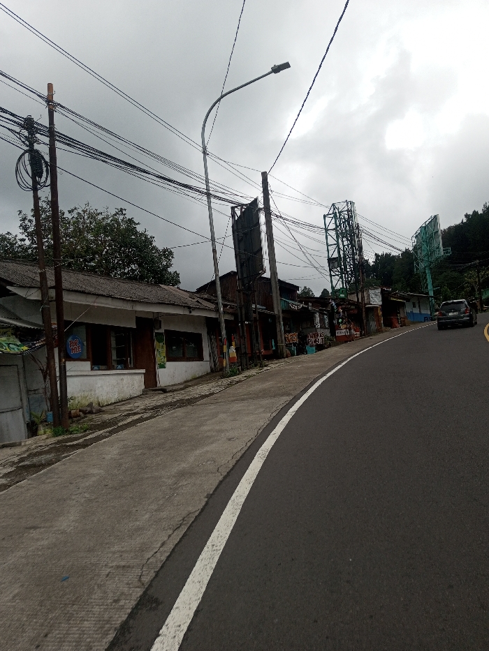 The image depicts a street scene in Bangtsho, Bhutan, under an overcast sky. The viewpoint is from a low angle, looking forward along an asphalt road that curves slightly to the right and descends. A solid white line delineates the left edge of the road, bordering a cracked concrete sidewalk.
On the left, a row of low-rise buildings lines the street. The nearest structure has white-painted lower walls, a dark weathered roof, multiple windows, a door, and a circular blue sign affixed to its facade. Further down the street, subsequent buildings exhibit varied materials, including corrugated metal, and some display commercial signage with text such as "MIE AYAM," "BASO RAHAYU," and "WARUNG RASA KITA." Multiple wooden utility poles, heavily strung with black electrical wires, and a metallic streetlight pole are prominent on this side. Further along, several large, empty green or dark metal billboard frames rise above the rooftops.
On the right side of the road in the mid-ground, a dark-colored SUV is visible. The background on the right is dominated by a densely forested hillside. No people are visible in the scene. The sky is uniformly grey and cloudy.