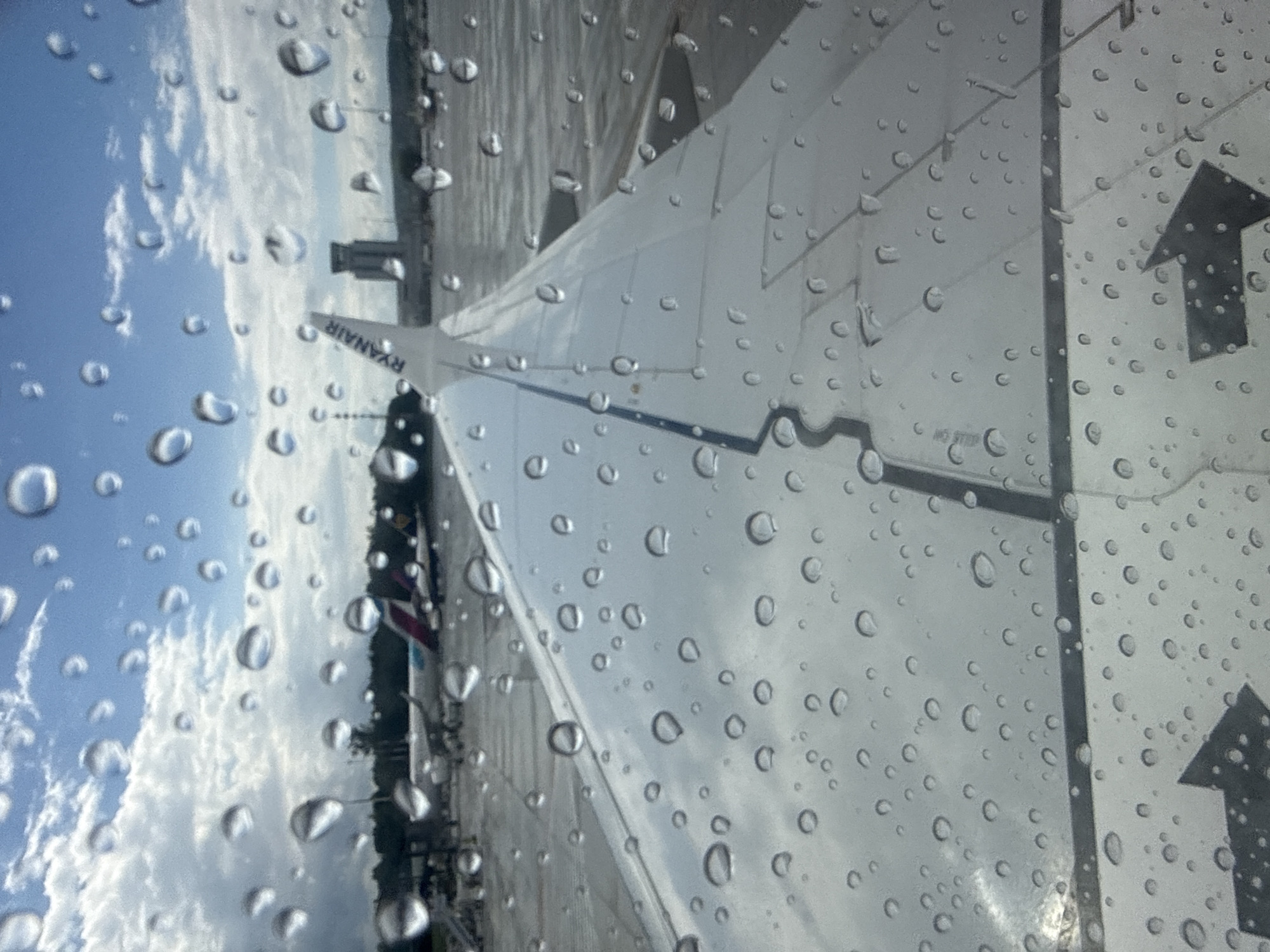 The image shows a close-up view of an airplane wing with water droplets on the window, taken from inside the aircraft. The wing is white with a dark stripe along its edge. The word "RYANAIR" is visible on the tail fin. In the background, there is an airport tarmac with other airplanes and airport buildings. The sky is cloudy with patches of blue. The scene appears to be at John Paul II International Airport Kraków–Balice, located in Balice, Poland. No people are visible in the image. The overall impression is a typical airport scene during or after rain.