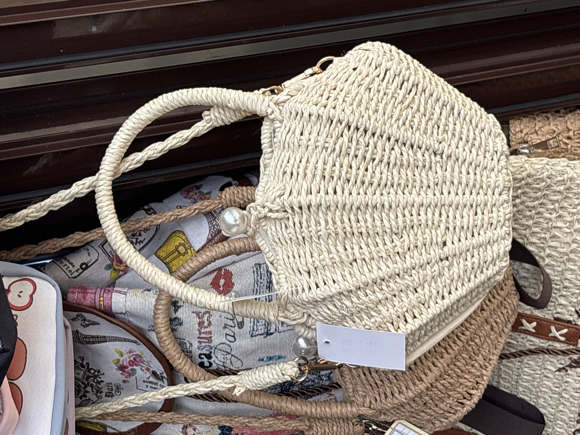 The image displays a close-up of several woven straw bags, with a prominent light-colored, hexagonal bag in the foreground. This bag is intricately woven with a thick, off-white cord and features a braided handle. Gold hardware connects the handle to the bag. A white tag with rounded corners is attached to the bag, and a pearl accent dangles from a strap near it. Beneath this bag, other woven bags are visible, including one in a natural straw color and another with a pattern that appears to include floral motifs, stylized buildings, and lipstick kisses. The background is a dark, reflective surface, possibly wood or a dark countertop, with horizontal lines suggesting drawers or shelves. The lighting suggests an indoor setting, possibly a market stall or shop, with no clear indication of time of day or weather. No specific emotions are conveyed. The visible text on the patterned fabric of the bag below includes "Paris" and what appears to be French text, such as "Banque de France". The location context of Sochi, Russia, does not provide specific visual cues within the image itself.