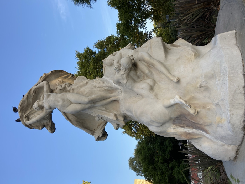 The image displays a large, weathered allegorical stone monument, prominently featuring a nude female figure ascending from a tumultuous base of other entangled figures, set against a clear blue sky. This sculpture is the Monumento a la Carta Magna y las Cuatro Regiones Argentinas, located at the intersection of Avenida del Libertador and Avenida Sarmiento in the Palermo neighborhood of Buenos Aires, Argentina.
The central female figure stands erect on an elevated part of the monument, facing slightly right, with her arms raised, holding aloft a voluminous piece of drapery that billows upwards and to her left. Her body is rendered with classical proportions. Below her, the rugged base of the monument is carved with multiple nude or semi-nude human figures in various dynamic poses, appearing to struggle or ascend. One figure on the right is partially submerged within the stone, with another smaller figure below, its head downturned, an arm extended. Traces of human forms, including a foot, are visible on the lower left side of the base. The light-colored stone exhibits natural discoloration and staining, particularly in the lower crevices, indicative of exposure to elements. Three dark-feathered birds are perched on the highest point of the drapery held by the central figure.
The monument is situated on a circular base. In the background, lush green trees frame the scene on the right and left, with a light-colored building partially visible behind the trees on the far left. The sky above is cloudless and a bright cerulean blue, suggesting daylight conditions.