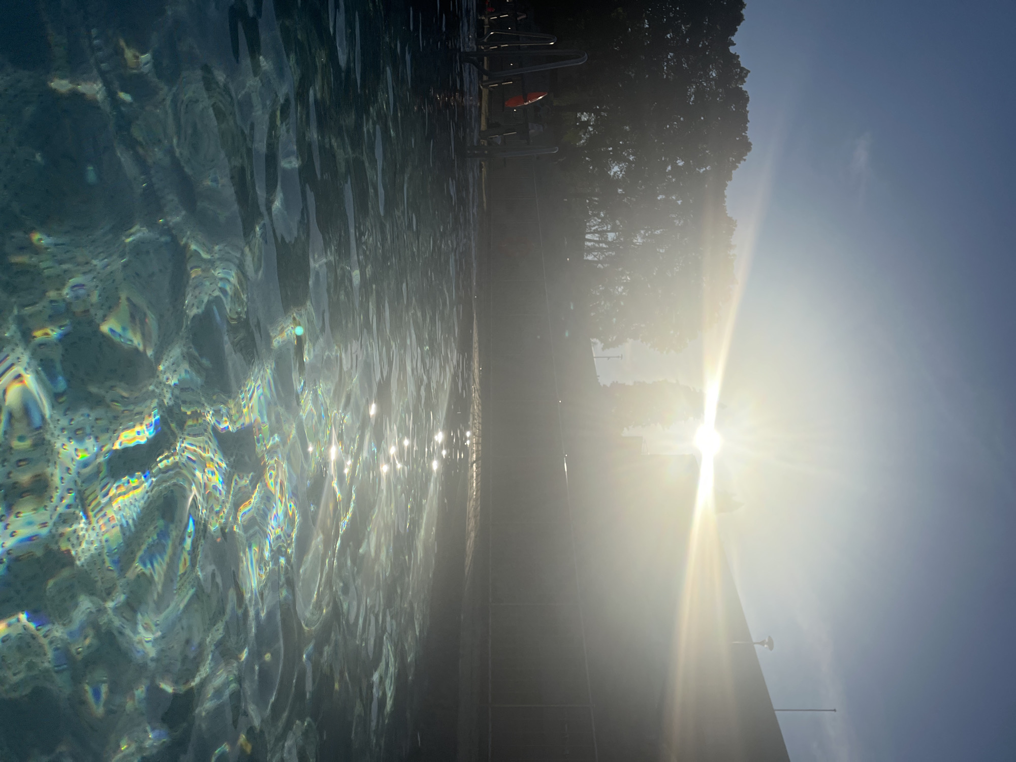 The image depicts a swimming pool during a bright, sunny day. The sun is very prominent in the sky, casting a strong glare and creating lens flares. The surface of the pool water is rippling, with sunlight reflecting off it in a dazzling display of colors, including rainbow hues. In the background, there are trees, a fence, and a building. There are also metal pool stairs and a life preserver visible by the edge of the pool. The overall impression is one of a peaceful, sun-drenched aquatic setting. The scene is captured from within the pool, looking towards the sun and the surrounding environment.