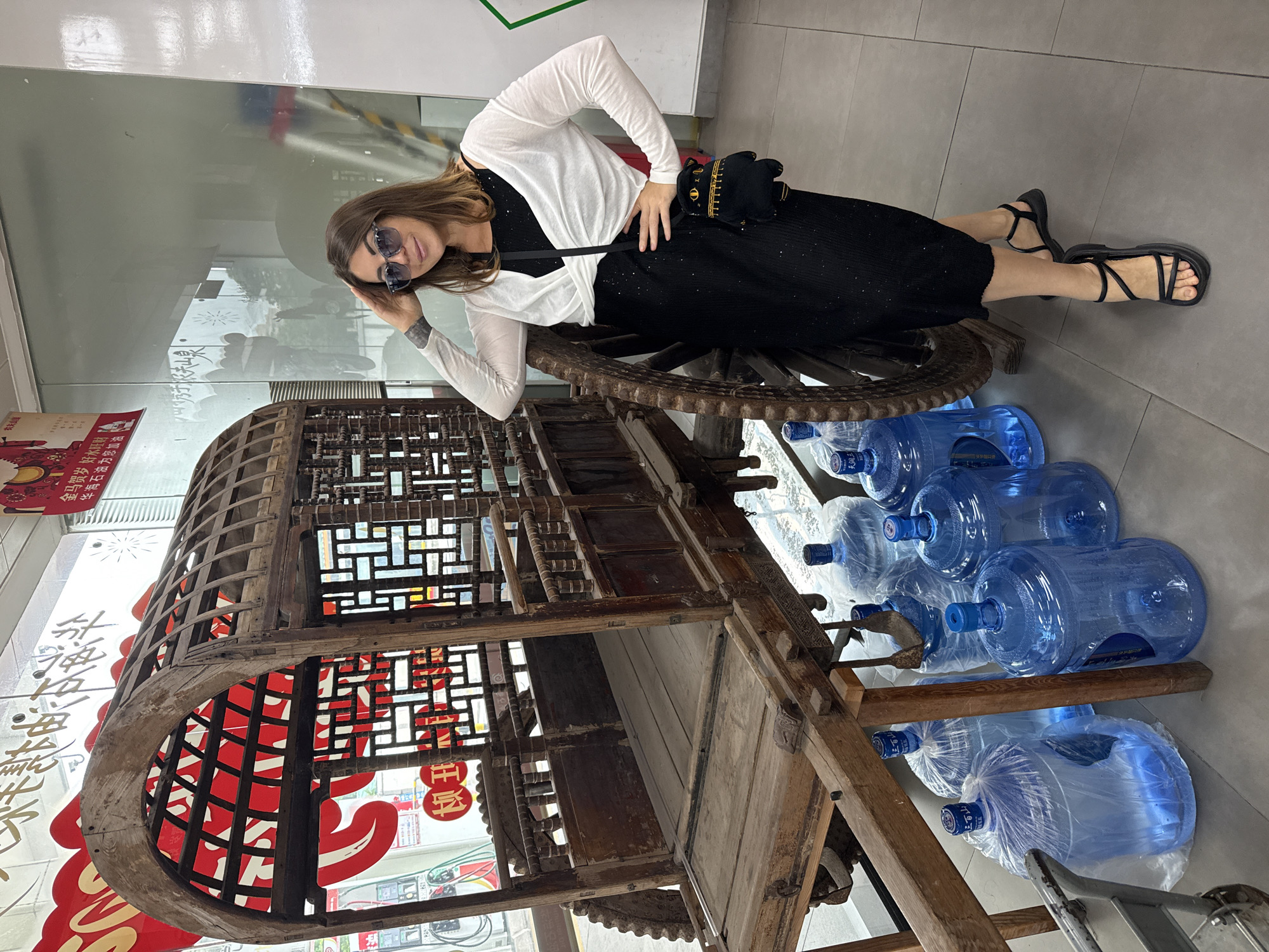 A woman stands next to an antique wooden cart in what appears to be a retail space in Huizhou, China. The cart is ornate, with a carved wooden frame and what looks like latticework on the upper portion. Behind the cart and the woman is a large glass window, likely a storefront, revealing some signage and what seems to be a service station or convenience store beyond. The woman is wearing a black sparkly dress, a white shrug, sunglasses, and black sandals. She has her hand on her head and is looking towards the camera with a smile. She is also carrying a small black purse shaped like a cat. In front of the cart and to the left of the woman are several large blue plastic water jugs, some of which are covered in plastic sheeting. The floor is tiled in a light gray color. The overall environment suggests a commercial or public indoor space.