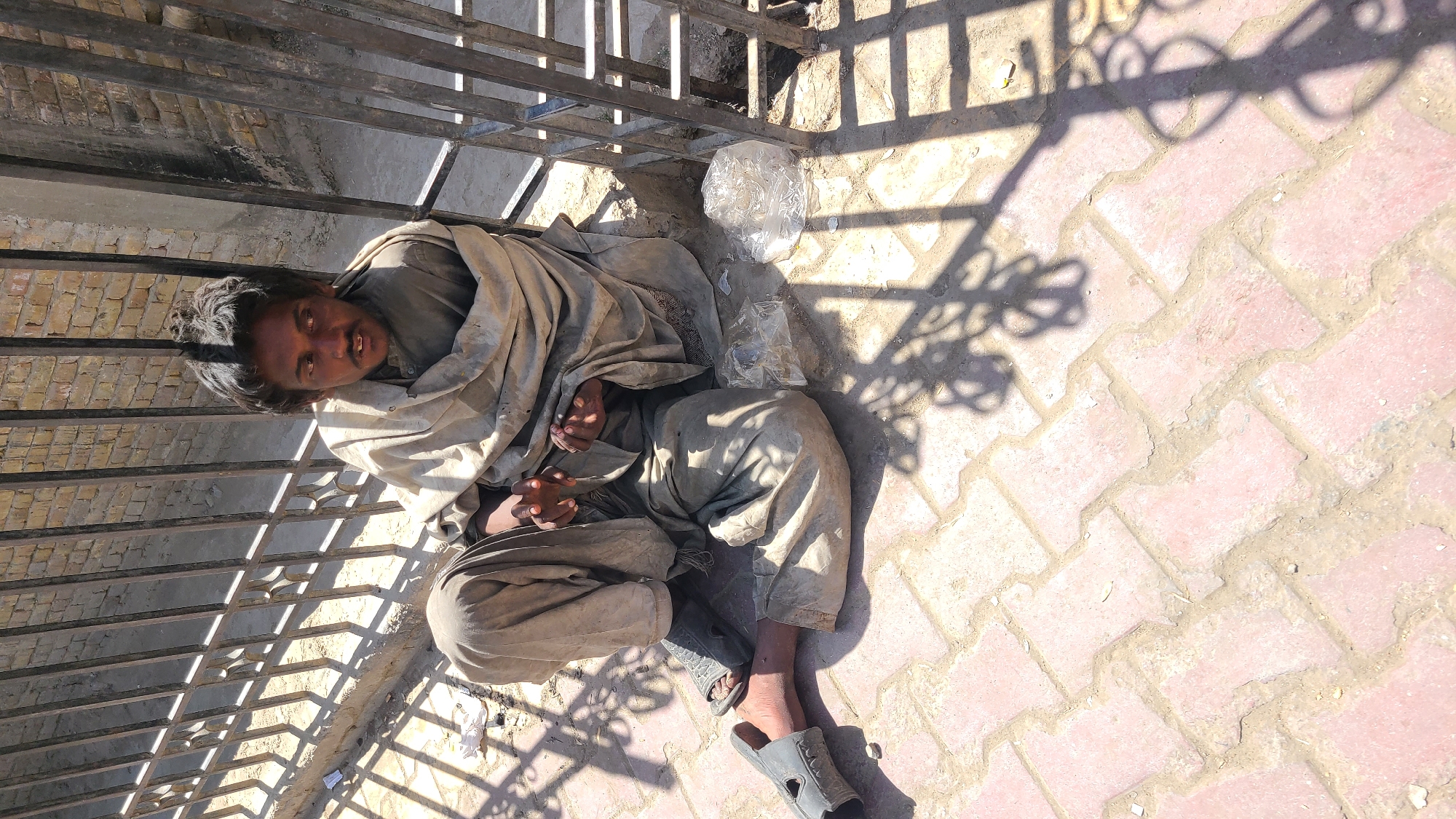 A man lies on a paved surface near a metal fence in Quetta City Tehsil, Pakistan. The man is dressed in soiled, light-colored clothing and is partially covered by a blanket. He has dark hair and appears to be looking up. Next to him are clear plastic bags. The area is illuminated by bright sunlight, casting shadows from the fence onto the pavement. The pavement is made of interlocking red and tan bricks.