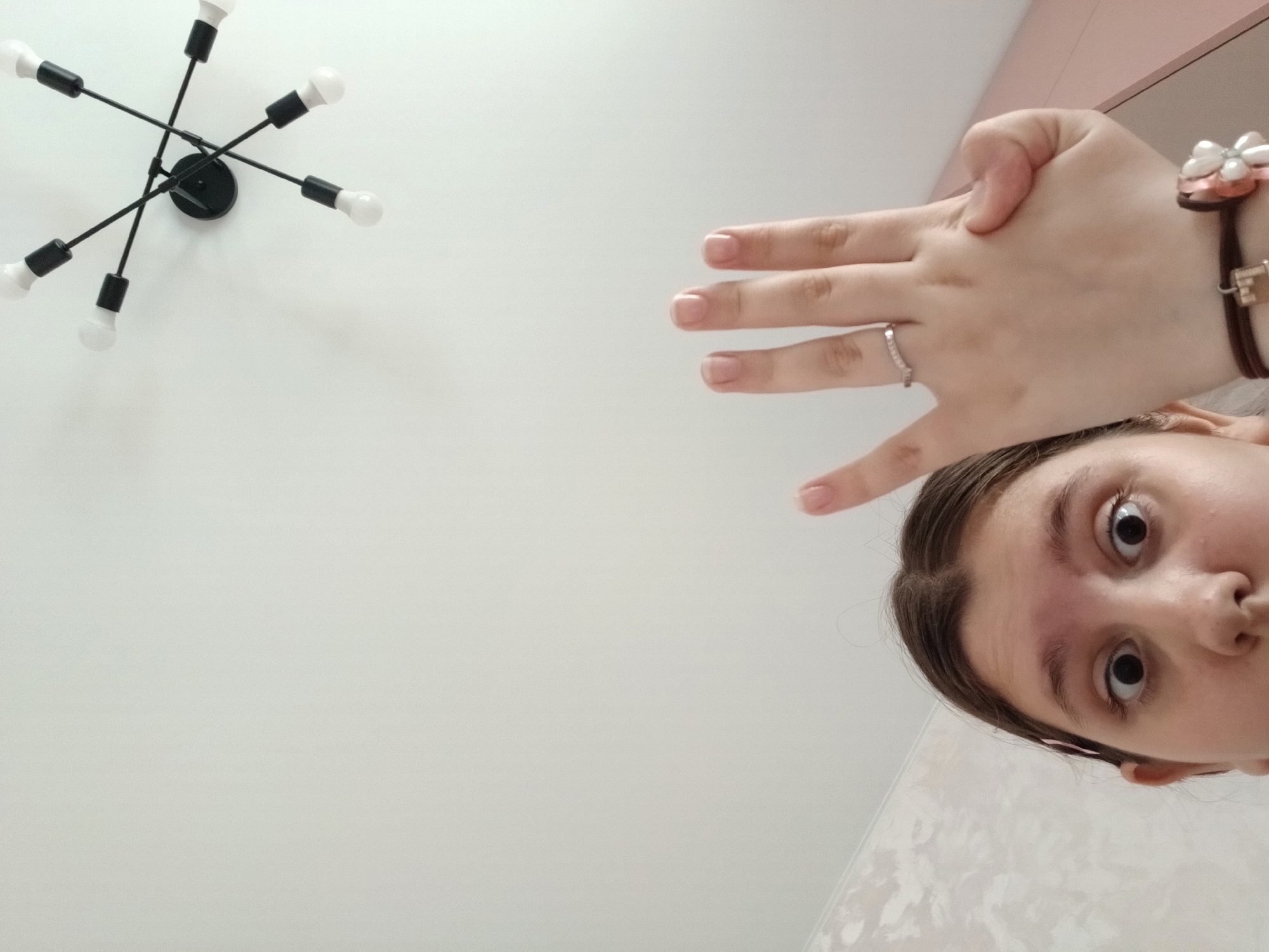 A photograph taken from a low angle depicts a ceiling with a light fixture, a hand, and a person's face. The light fixture is a modern design with multiple arms extending from a central point, each ending in a light bulb. A hand with a ring on one finger is positioned near the light fixture. A person's face, partially visible, is situated near the hand. The background is a plain white ceiling. The location of the image is Марковский, Russia.