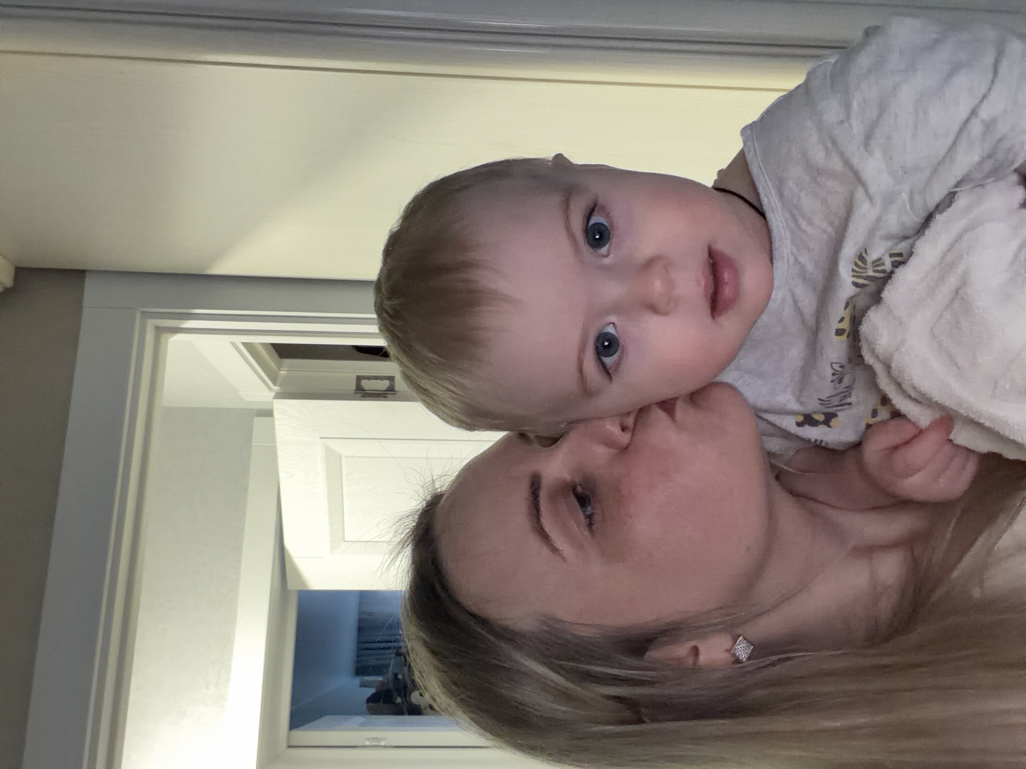 The image depicts an indoor domestic scene featuring an adult female interacting with an infant. The setting consists of light-colored walls and doorframes, with no visual information within the frame to confirm a specific geographical location, such as Votkinsk, Russia.
The adult female, positioned on the left, has long, light-colored hair and is wearing a light-toned garment. She has a square, possibly diamante, stud earring visible on her left earlobe. Her head is angled towards the infant's left cheek, where she is engaged in kissing. Her eyes appear closed or cast downwards in profile.
The infant, held by the female, is positioned centrally and slightly to the right, facing directly towards the viewer. The infant has light blonde hair, blue eyes, and an open mouth. The infant is wearing a light grey long-sleeve top with faint, patterned details, and is partially wrapped or covered by a white towel or blanket.
In the background to the left, a doorway is visible, leading to an interior room that appears darker with discernible blue textile elements, possibly curtains. To the right, another section of a light-colored wall and doorframe is visible. The lighting is artificial and uniform.