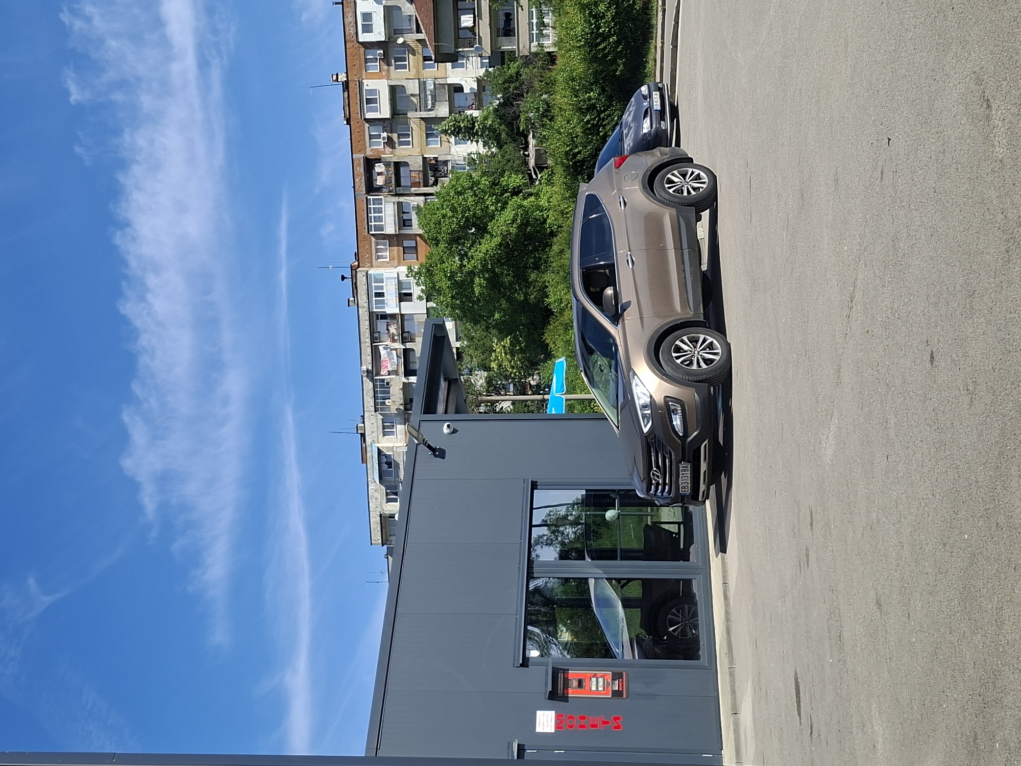 The image depicts an outdoor scene in Unknown, Unknown, featuring a building with "MONEYTS" signage, a parked brown Hyundai Santa Fe with license plate EB3131BA, and another dark-colored car parked further away. A multi-story apartment building rises in the background amidst lush green trees, set against a clear blue sky adorned with wispy white clouds. The setting appears to be a parking area adjacent to the building, marked by gray pavement.