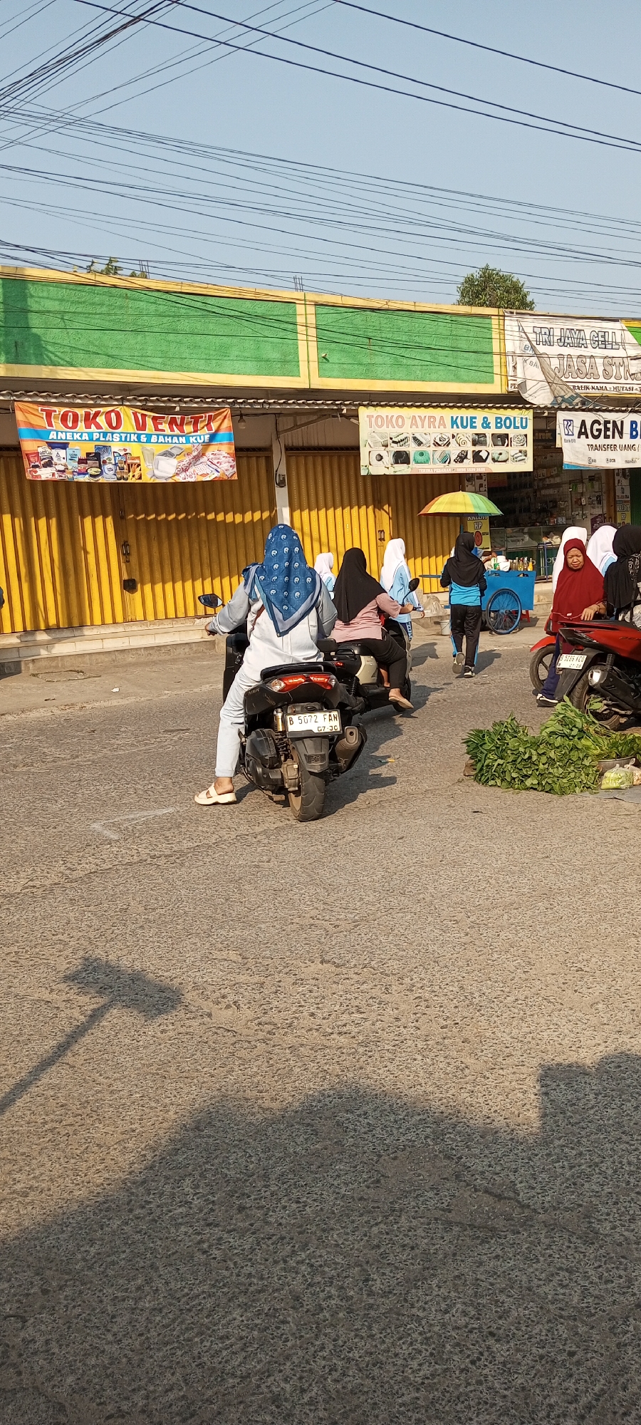 A street scene unfolds in Setu, Indonesia, under a bright, cloudless sky. In the foreground, the textured asphalt road shows a distinct shadow of a street lamp or pole stretching across its surface. The focal point is a line of motorcycles and pedestrians navigating the road. A black motorcycle with the license plate "B 5072 FAN" is prominent, carrying two individuals; the rider wears a blue patterned headscarf and a light-colored outfit, while the passenger is also veiled. Behind them, two more motorcycles follow, each with veiled occupants.
To the right, a pile of fresh green vegetables sits on the roadside, suggesting a local market or vendor. Further down the street, a blue cart with a colorful umbrella is visible, and several individuals are interacting near shops. The shops themselves are characterized by bright yellow rolling metal doors, partially open to reveal merchandise and signage. One banner overhead reads "TOKO VENTI" with images of various products, and another proclaims "TOKO AYRA KUE & BOLU" showcasing an assortment of cakes. Above the shops, a green facade with yellow trim is visible, and a network of power lines crisscrosses the sky. The overall impression is one of a busy, sunlit day in a commercial area of Setu, Indonesia.
