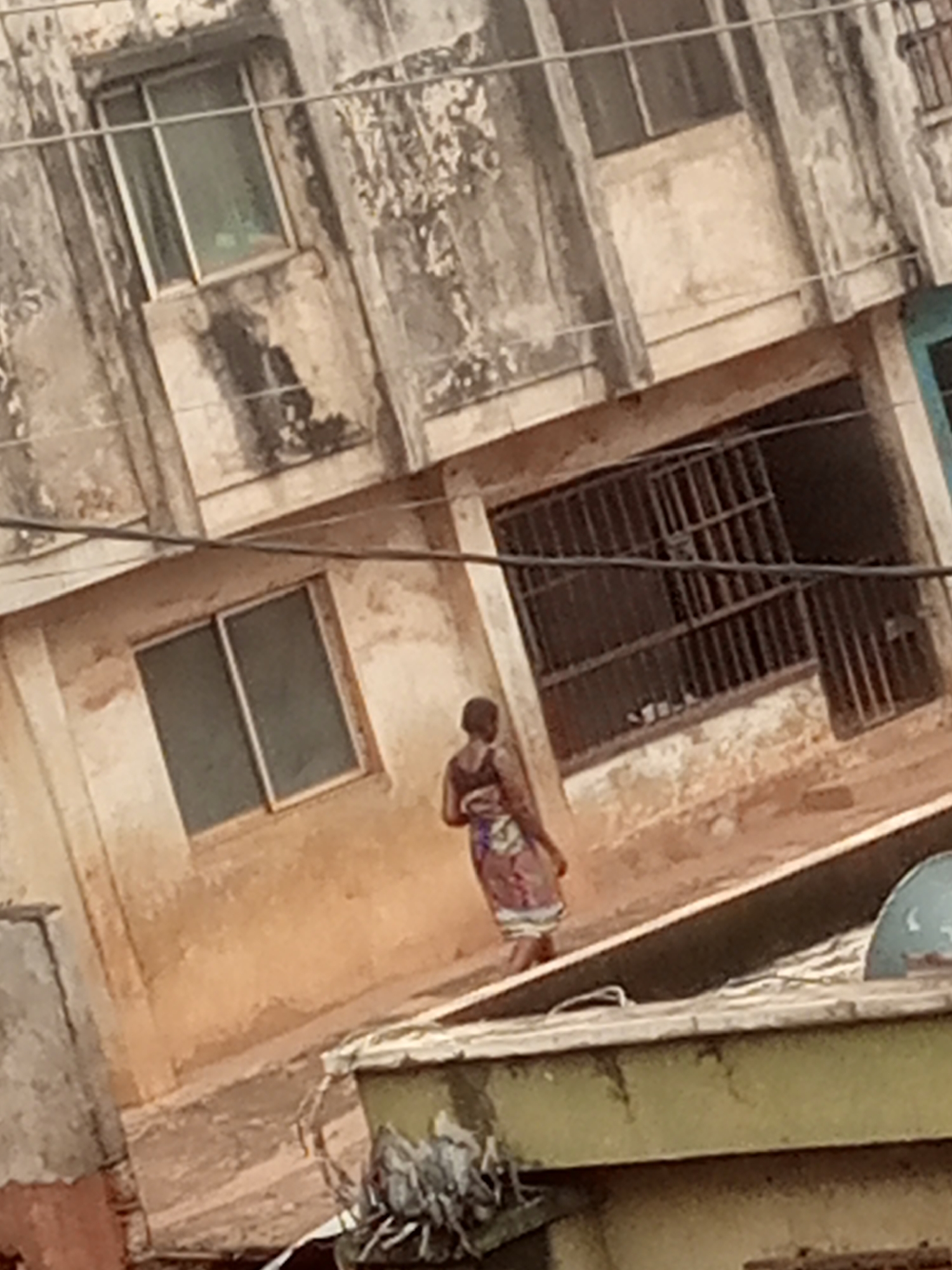 An outdoor, medium-angle shot shows a woman wearing a patterned dress and carrying a bundle on her back. She is walking towards the right side of the frame in front of a dilapidated building. The building has multiple windows, some with bars, and appears to be made of concrete and plaster, with visible signs of age and weathering. The ground in front of the building is a reddish-brown color. In the foreground, there is a ledge with some debris on it, and a circular object that appears to be a blue cap. The image is slightly blurry and tilted.