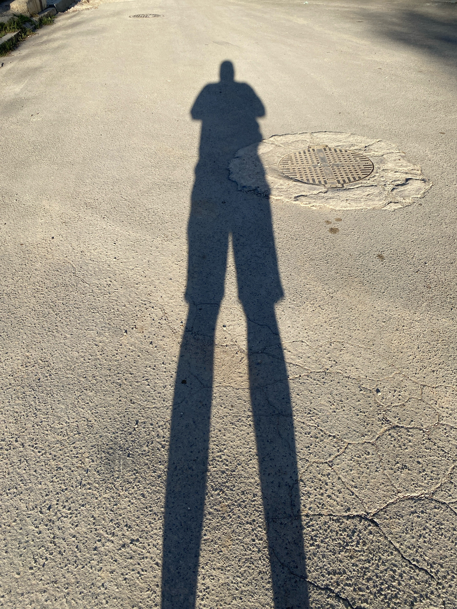 The image depicts a long shadow of a person standing on a cracked asphalt surface. The shadow, cast by a low sun, stretches diagonally across the frame from left to right. A round manhole cover with a grid pattern is visible on the right side of the asphalt. The environment suggests an outdoor urban setting, possibly a street or sidewalk in Chișinău, Moldova, characterized by the worn asphalt and a visible drainage grate. The overall lighting indicates a sunny day.