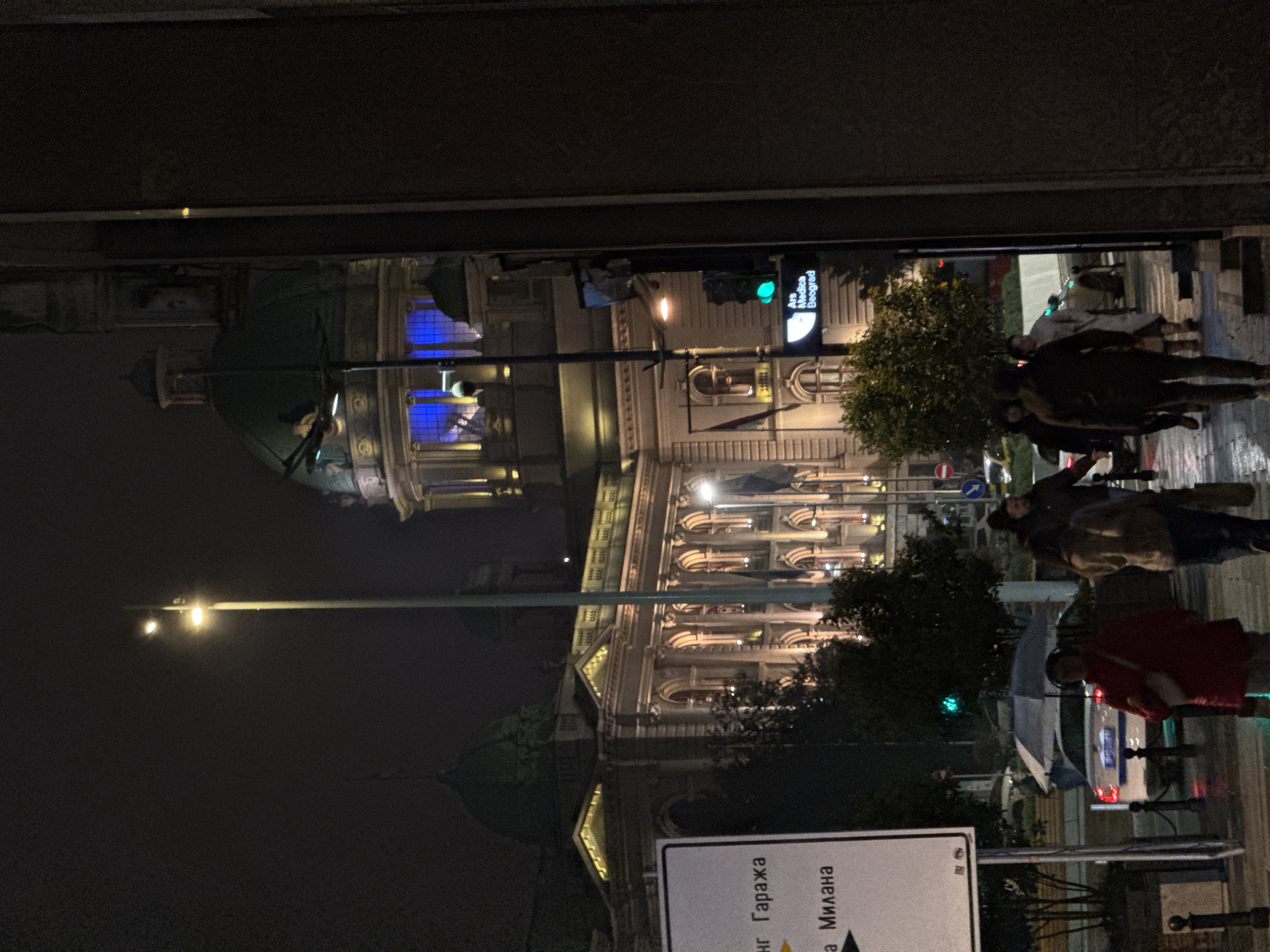 The image shows a night scene in Belgrade, Serbia, featuring the National Assembly building illuminated in the background. A street lamp is visible, and the street appears wet, possibly due to rain. Several people are walking on the sidewalk, some carrying umbrellas. A street sign is visible, with text in Serbian, including "Гаража" and "Милана". A traffic light is also present, showing green. A building with architectural detail and blue lights is visible behind the trees.