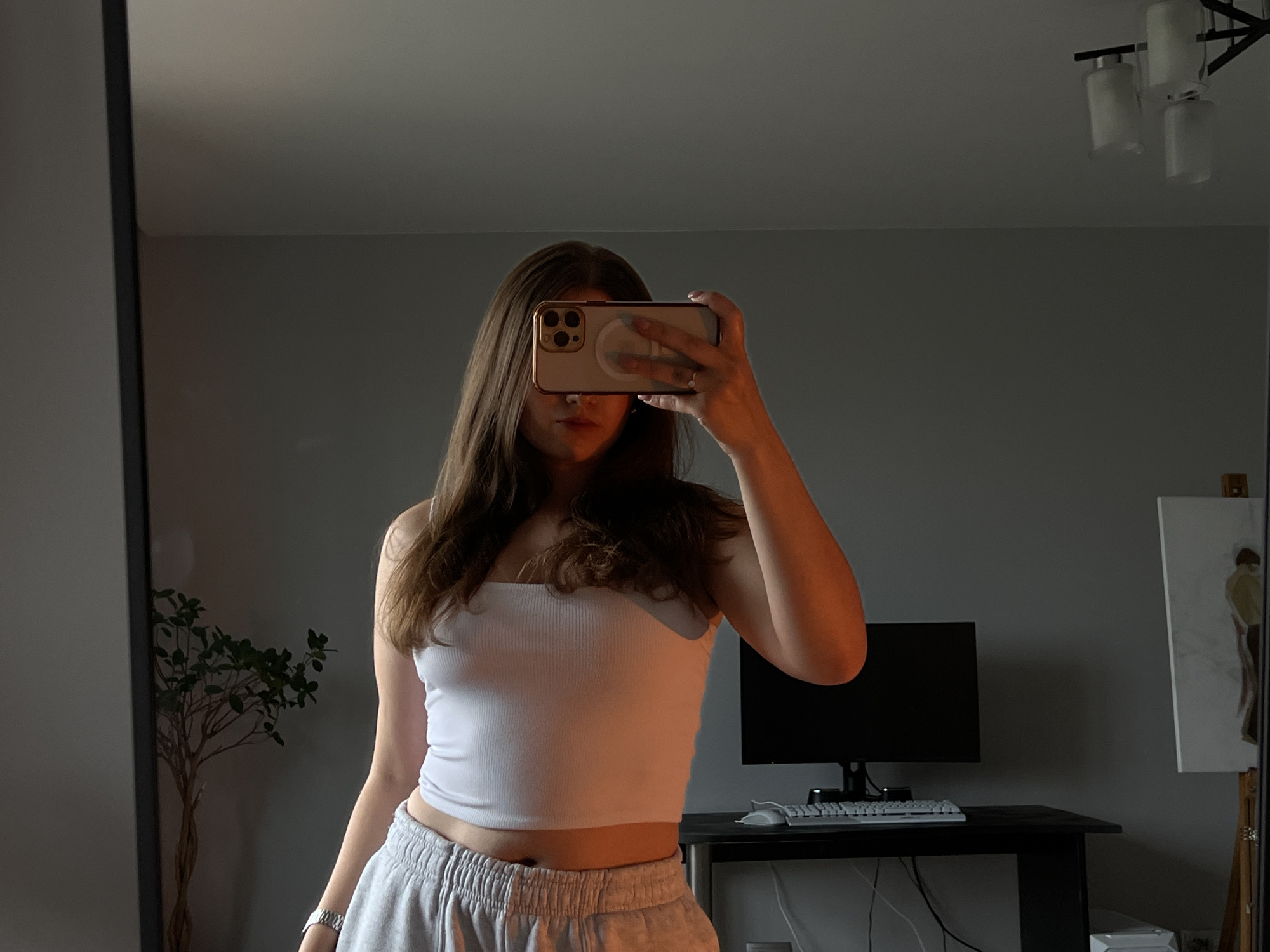 A young adult female is depicted in a mirror selfie, standing indoors in a room located in Ternopil, Ukraine. Her face is obscured by a smartphone, held horizontally in her right hand, which is equipped with a light-colored case featuring a gold border and a clear circular MagSafe ring on the back. A ring is visible on her right ring finger. She wears a white ribbed tube top and light grey athletic sweatpants with an elasticated waistband, revealing her midriff. A silver-toned watch is on her left wrist.
The scene is illuminated by a warm, orange-toned light source, casting a glow on her skin, clothing, and parts of her hair. Her long, brown hair falls over her shoulders. The room features plain, light grey walls and a matching ceiling. In the background, to the left of the subject, a dark vertical mirror frame is visible, alongside a small, leafy, tree-like plant with multiple intertwined trunks. Above, a ceiling light fixture with a dark metal frame and three white cylindrical shades is mounted. To the right, a black desk holds a black computer monitor, a white keyboard, and a white mouse. Further to the right, a wooden easel displays a white canvas with a minimalist drawing of a standing figure in earthy tones.