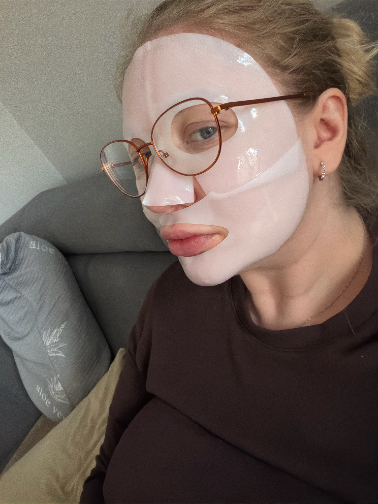 A woman is shown in a close-up, selfie-style shot. She is wearing a pale pink sheet mask that covers most of her face, with cutouts for her eyes, nose, and mouth. She has on round, wire-rimmed glasses and a small hoop earring in her visible ear. Her hair is pulled back, and she is wearing a dark brown crewneck sweatshirt. The background suggests an indoor setting, possibly a living room, with a grey couch and pillows. One pillow on the left has the text "aloe ve" visible. The image is taken from a slightly upward angle. There is no indication of the city of Koszalin, Poland in the image.