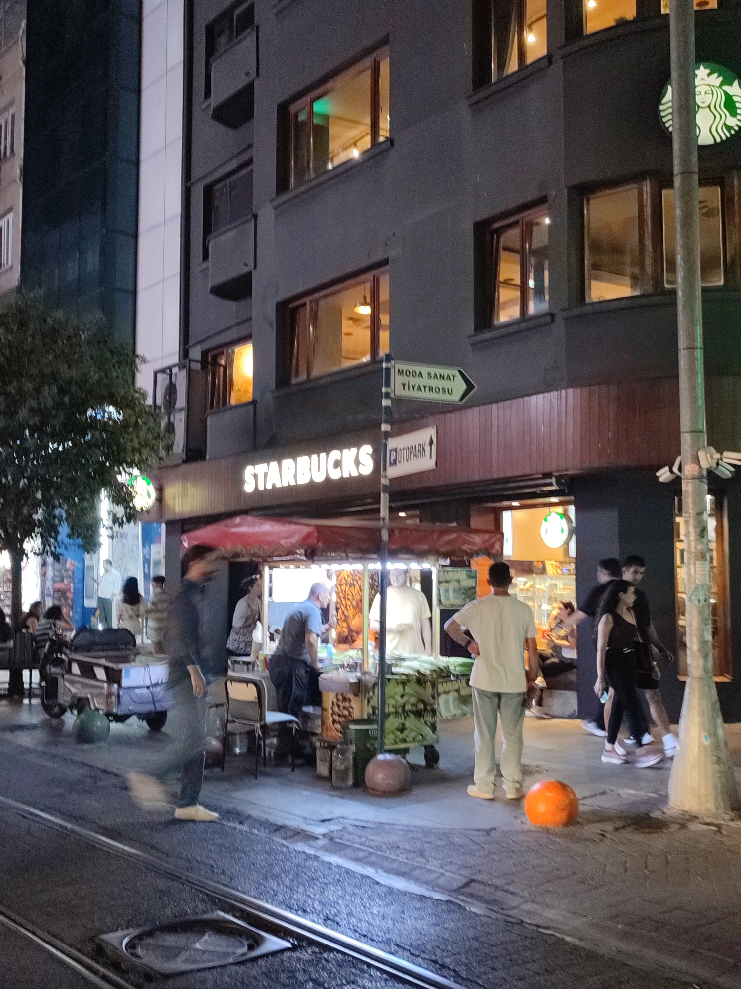 The image depicts a street-level view at night in Kadıköy, Turkey. A Starbucks coffee shop is prominently featured with its illuminated signage. Adjacent to the Starbucks, a food cart is visible, staffed by at least one individual serving customers. Several people are gathered around the food cart and the Starbucks entrance, including individuals standing, walking, and seated. A man in a grey t-shirt and dark pants is standing at the food cart, and another man in a white t-shirt and light-colored pants stands facing away from the camera. A woman in a black tank top and shorts is seen walking with another person. In the foreground, a streetcar track is visible on the asphalt road, and an orange spherical object rests on the sidewalk. A light pole with security cameras is located on the right side of the frame. The building housing the Starbucks has multiple windows, some illuminated from within. A green directional sign pointing to "MODA SANAT TİYATROSU" (Moda Art Theater) is mounted on a pole. The overall impression is of a busy urban commercial area at night.