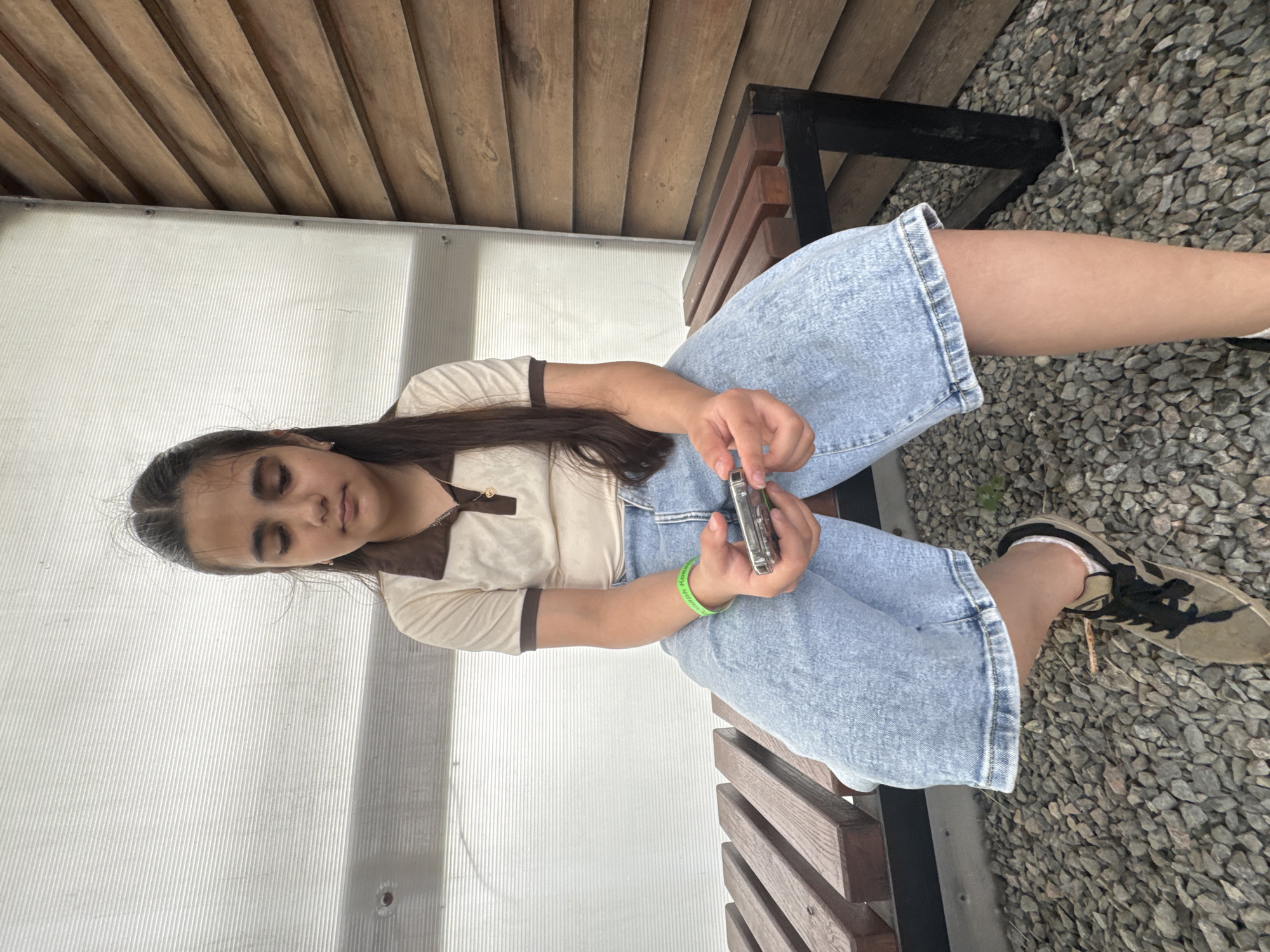 A young girl is sitting on a wooden bench in Zaliznychne, Ukraine. The bench is outdoors, with a pebbled ground and a wooden wall behind it. The girl is wearing a light brown polo shirt with a dark brown collar and trim, and light blue wide-leg jeans. She is holding a small silver object, possibly a phone or music player, in her hands. She has long dark hair and is looking down at the object. A green bracelet is visible on her left wrist. The background consists of a white translucent wall on the left and a horizontally slatted wooden wall on the right.