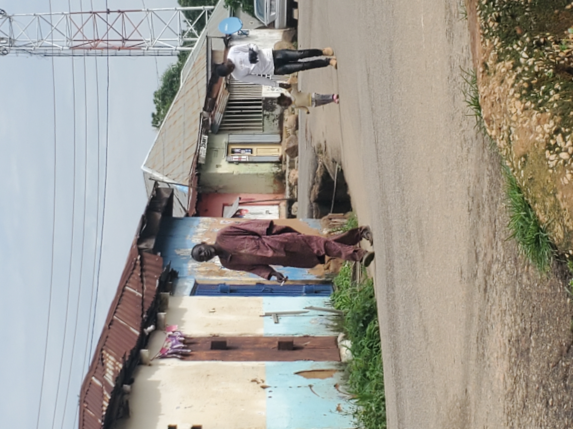 The image shows a street scene in Jos, Nigeria. In the foreground, a man in a maroon traditional outfit stands on the road, holding a cigarette. Behind him, a building with a rusted corrugated iron roof and a blue door is visible. Further back, another man in a white shirt and black pants is walking with a child dressed in a yellow top and grey pants. The background features a large metal tower structure and some trees. The sky is overcast, suggesting cloudy weather. The scene appears to be an outdoor, urban environment. There is no visible text in the image.