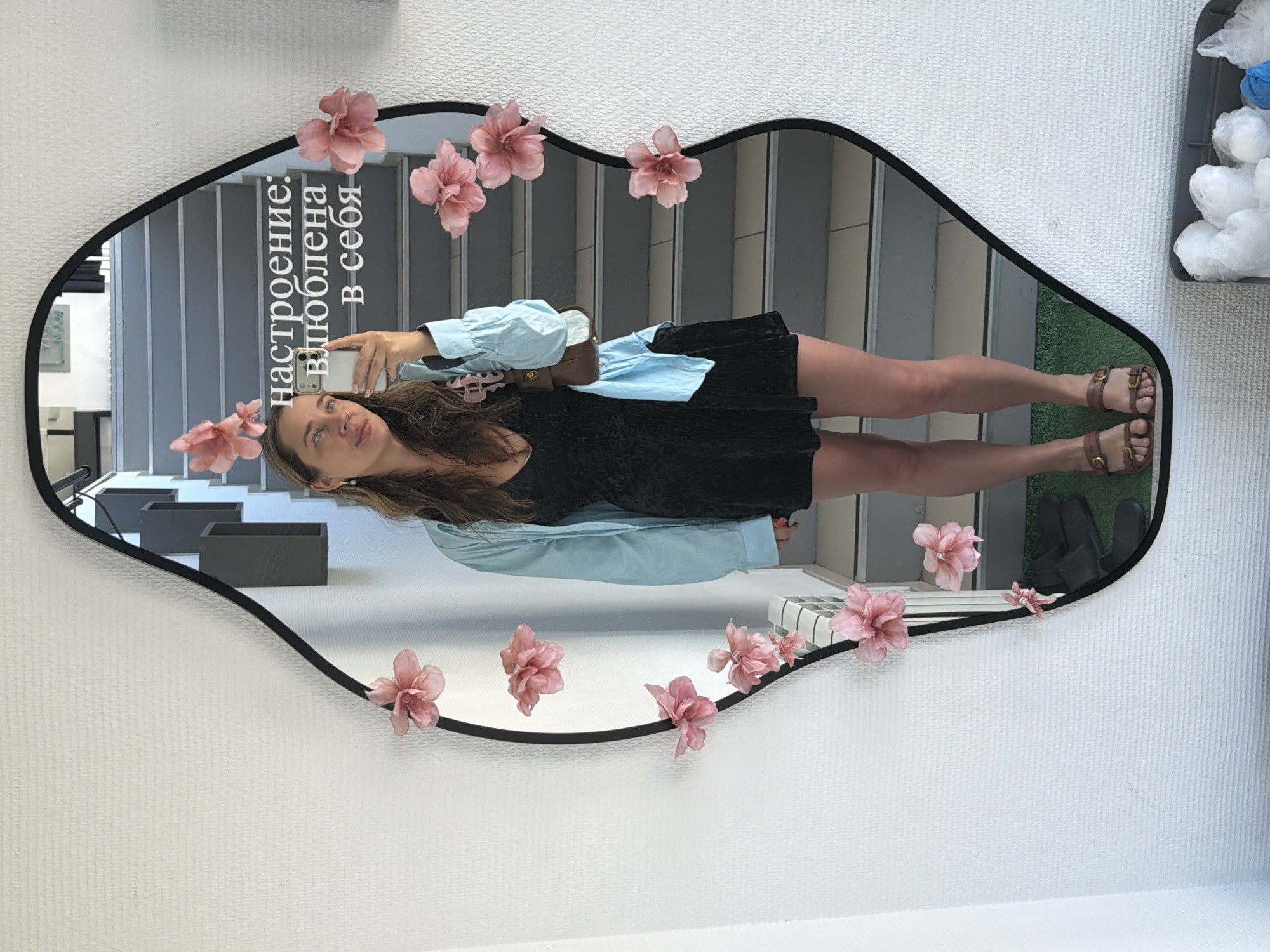 A full-body mirror selfie shows a woman standing and looking towards the camera with a slight smile. The mirror has a distinctive irregular, lip-like black frame decorated with multiple artificial pink flowers. The woman wears a black top or dress, a light blue long-sleeved shirt, and brown buckled sandals. She holds a white smartphone in her hand and carries a small brown purse across her body.
The reflection in the mirror reveals an indoor environment, likely in Chaikovsky, Russia. Behind the woman, a set of gray and white stairs or steps is visible. To the right of her feet, artificial green turf covers the floor, with a pair of dark shoes or slippers resting on it. In the upper right of the reflection, a container holds crumpled white objects, possibly plastic bags, and a blue item. Small black rectangular planters are also visible near the bottom left of the reflection. The wall on which the mirror hangs is white and textured. White text is visible on the mirror's surface, reading in Russian: "настроение: влюблена в себя," which translates to "mood: in love with myself."