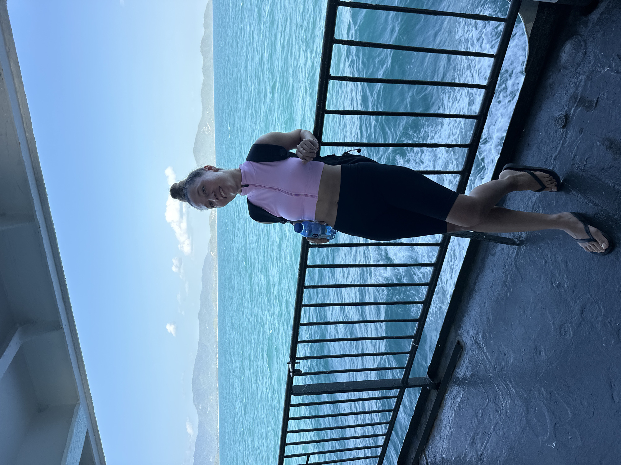 A young woman with her hair in a bun is standing on the deck of a ferry, leaning on the railing, with a bottle of water in her hand. She is wearing a pink crop top and black biker shorts, with a black backpack slung over one shoulder. She is smiling at the camera. The ferry is moving through turquoise water, with a mountainous coastline in the background. The sky is clear blue with a few white clouds. The scene appears to be in an island location in Thailand.