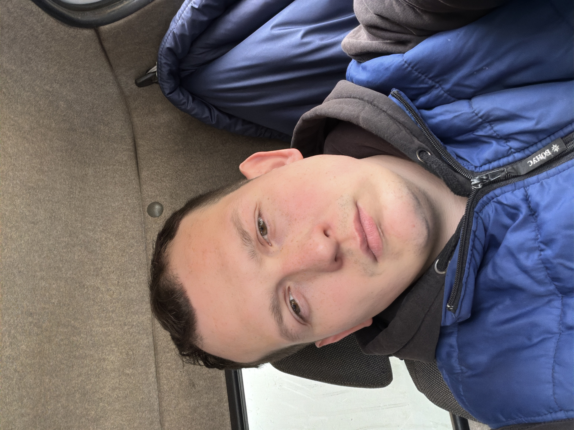 The original media file is a portrait-oriented photograph, depicting a young man in what appears to be an indoor setting, possibly inside a vehicle, rotated 90 degrees clockwise from its natural upright position. The man is looking directly at the camera, suggesting a selfie.
**Subjects:** The primary subject is a young man with fair skin, dark hair, and brown eyes. He is wearing a blue puffer jacket unzipped over a dark gray or black hooded sweatshirt.
**Scene and Setting:** The background consists of a light beige, textured material, which could be the interior lining of a car, bus, or train. To the man's left, a dark blue rolled-up item, possibly a blanket or pillow, is visible. A dark rectangular element, possibly a window frame or part of a seat, is seen to his right.
**Activity:** The man is posing for the camera, with a neutral to slightly contemplative expression.
**Notable Details:** The man has faint stubble and some freckles on his nose and cheeks. On the zipper pull of his blue jacket, the Cyrillic text "BOHYC" is visible, accompanied by a snowflake icon. The lighting appears to be ambient and even, without strong shadows, making the time of day undeterminable. There are no direct visual cues in the image itself to confirm the location as Chaikovsky, Russia, other than the provided context.