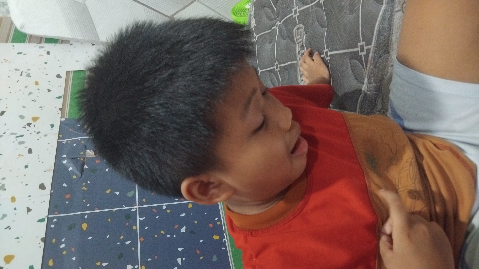 The image shows a close-up, overhead view of a young boy lying down, possibly sleeping or resting. He has dark, spiky hair and is wearing an orange t-shirt. His eyes are closed, and his mouth is slightly open, with his tongue visible. His right hand is visible, with a finger pointing towards the boy's shirt. The boy is lying on a surface that appears to be a mattress with a patterned fabric. The background shows a tiled floor with a colorful, speckled design, and a portion of a white piece of furniture. The lighting suggests it is daytime. The boy's expression and posture imply a state of rest or sleep. The overall impression is an intimate, everyday moment. There is no visible text in the image. The location context is Bangun Rejo, Indonesia.