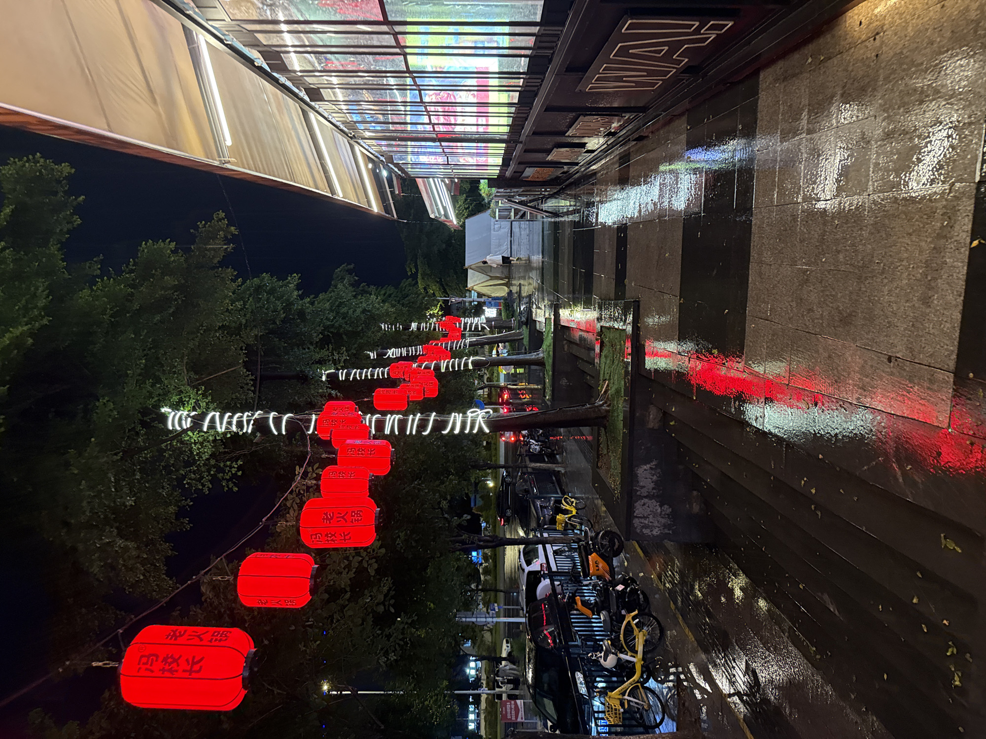 This image depicts a night scene in Haizhu District, China, possibly after a rain, as indicated by the wet, reflective ground. The perspective is rotated 90 degrees counter-clockwise.
The scene features a street lined with trees on the left, adorned with strings of white lights and numerous red, cylindrical lanterns. These lanterns, some with visible Chinese characters, create a festive and traditional atmosphere. On the right, a building with a dark facade runs along the street. Above, a section of the building's roof appears to be a glass canopy displaying colorful, illuminated signage, likely for shops or restaurants. The ground in front of the building is paved with large, dark tiles, some of which are checkered, and is wet, reflecting the red and white lights from the street and the building above.
Several parked vehicles, including cars and bicycles (some appearing to be shared bikes), are visible along the right side of the street. There are no clearly identifiable people engaged in specific activities, though the presence of the parked vehicles suggests a public or commercial area. The overall mood is atmospheric, with the artificial lights casting reflections on the wet surfaces, hinting at a possibly cool or damp evening. The visible text on the lanterns is in Chinese characters.