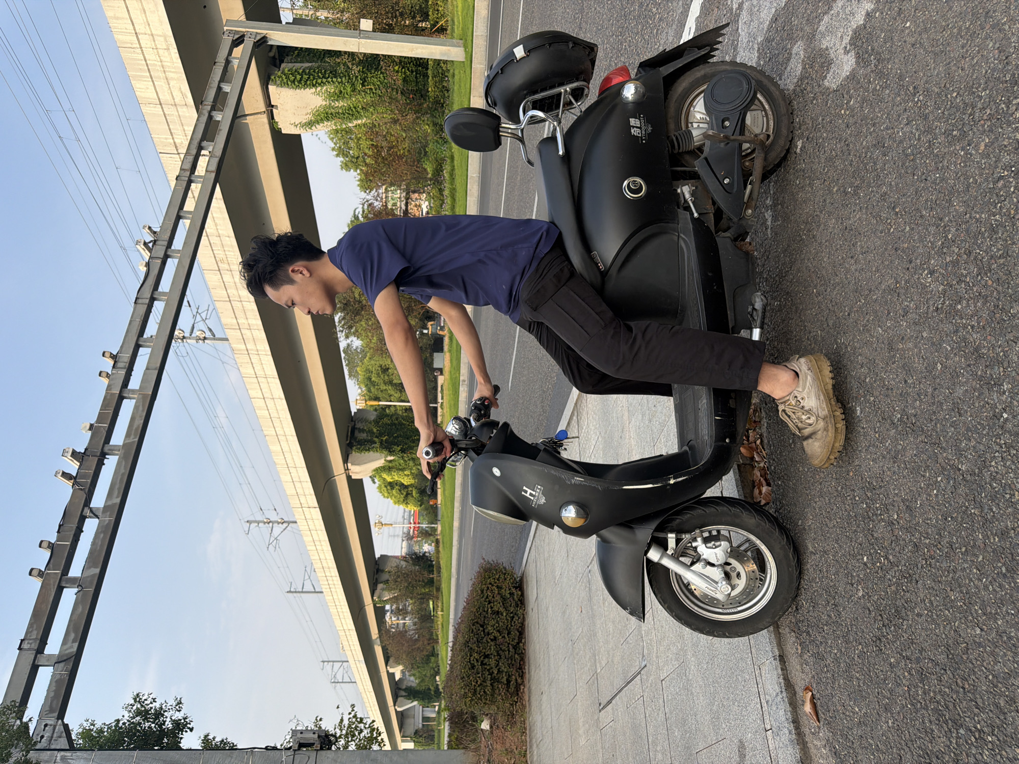 A young Asian male sits on a black electric scooter on a paved street in Jiangjin, China. He is wearing a dark blue t-shirt and black pants. The scooter has a storage box mounted on the rear. In the background, an elevated train track structure is visible, along with trees and a blue sky. The street is bordered by a sidewalk with some fallen leaves.