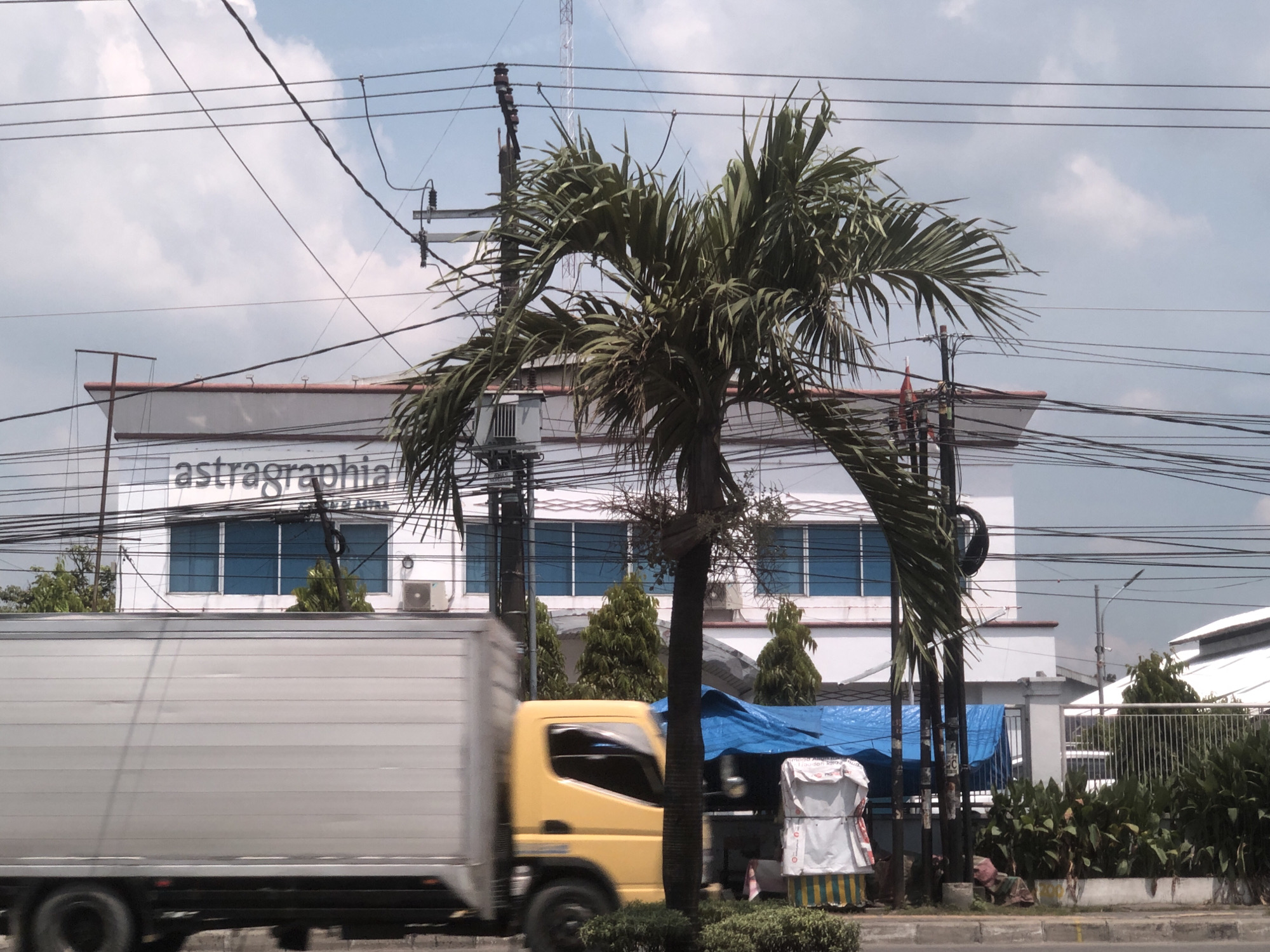 The image depicts a street scene in Medan, Indonesia. A yellow and white delivery truck is moving left to right in the foreground, blurring due to its motion. A palm tree is centrally located, obscuring part of a two-story white building with the word "astraphia" on it. Blue-tinted windows are visible on the building's facade. Overhead, a dense network of power lines crisscrosses the sky. Further in the background, a communications tower is visible. A blue tarp covers some items along the roadside, and a small white and yellow striped structure is also visible. Green shrubs border the road. The sky is partly cloudy.