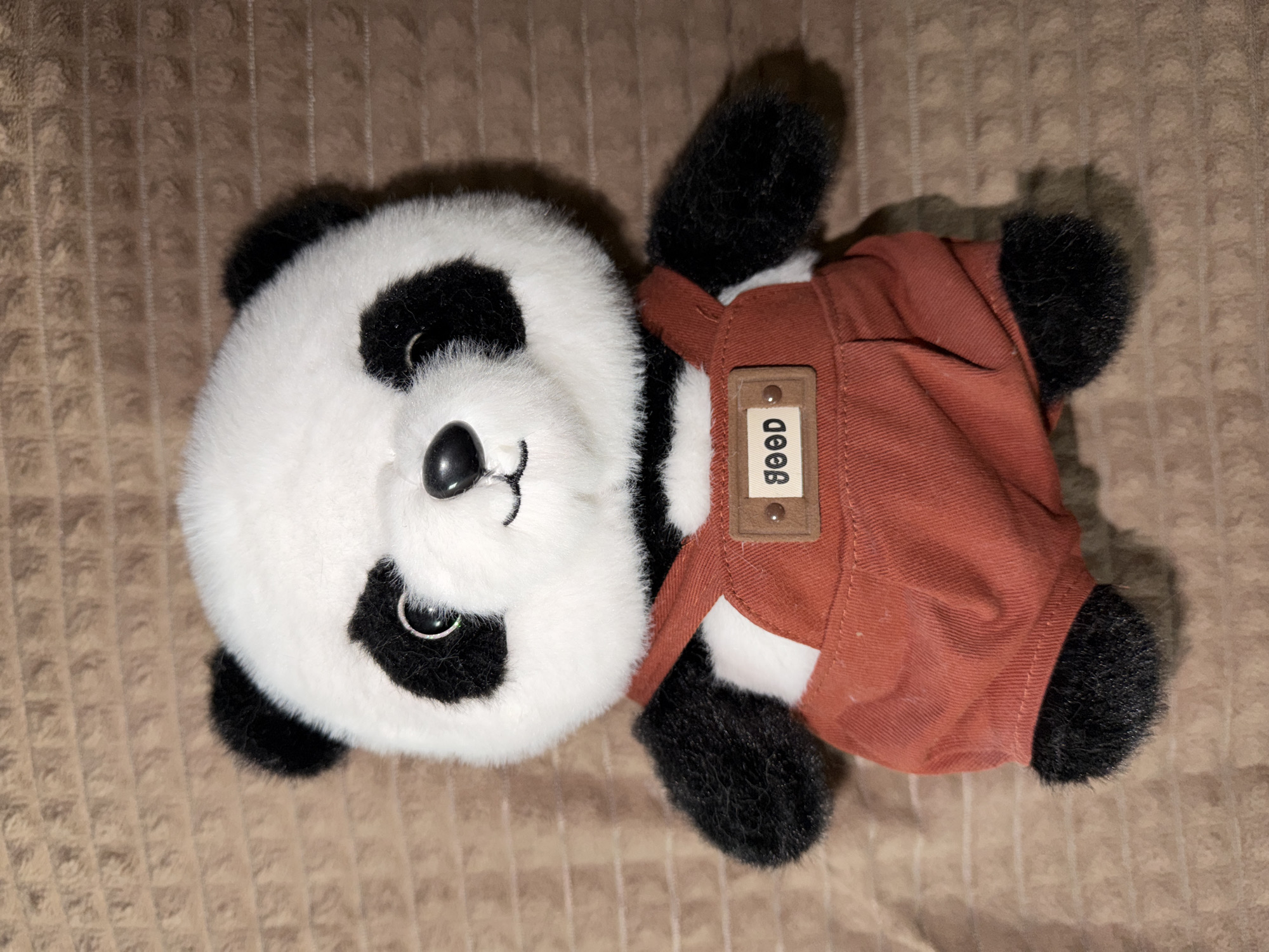 A plush toy panda is displayed from the chest up. The panda has white fur with black markings around its eyes and ears. It wears a pair of rust-colored overalls with a small brown tag on the bib that reads "GOOD". The panda is positioned centrally on a textured brown fabric background. There are no people or specific locations within the city of Naberezhnye Chelny, Russia visible in this image.