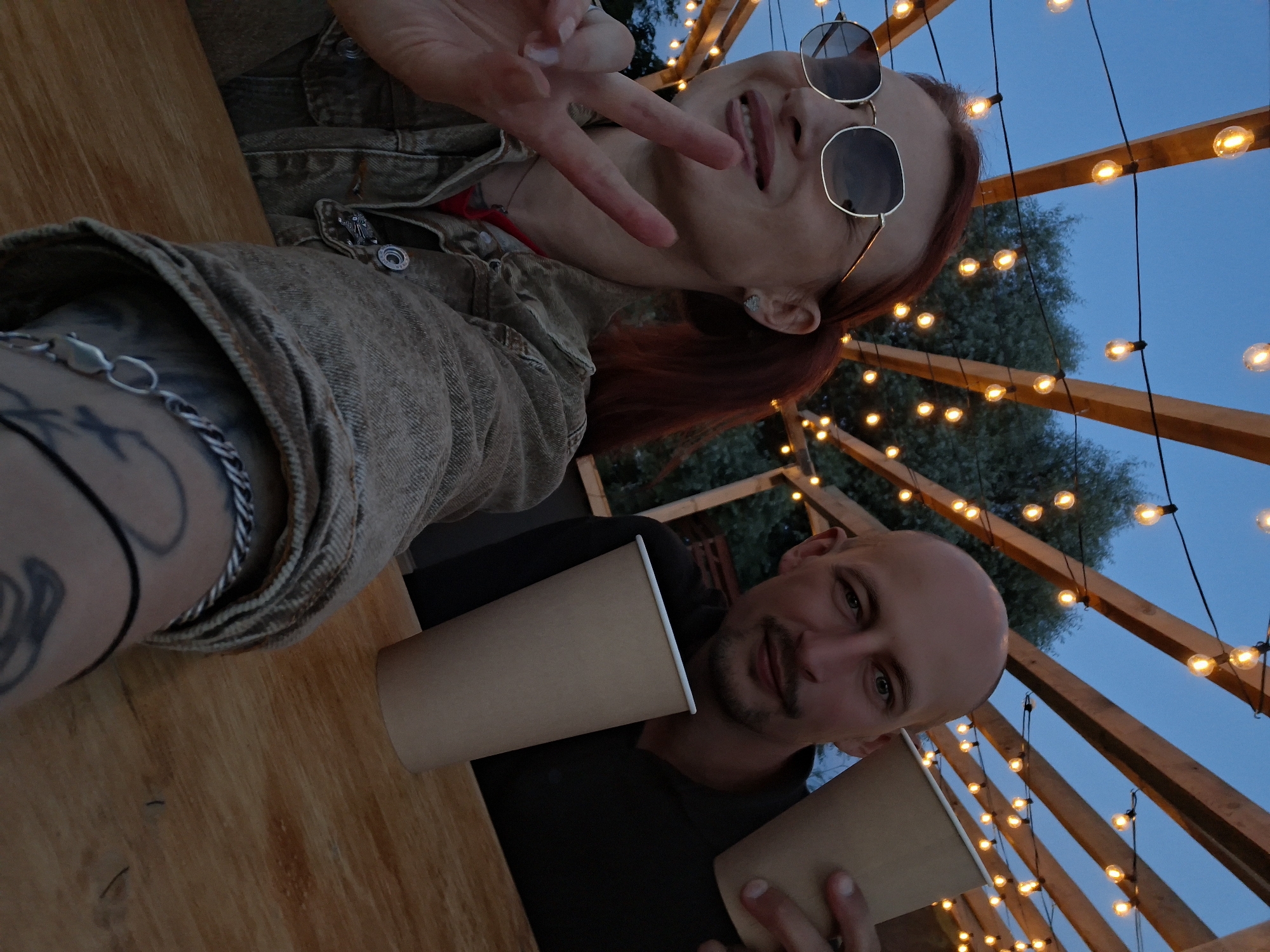 A man and a woman are taking a selfie at dusk, outdoors. They are seated at a wooden table under a pergola decorated with string lights. The woman, on the left, has red hair, wears sunglasses, and a denim jacket. She is holding up two fingers in a peace sign. The man, on the right, is bald with a mustache and is holding a disposable cup. Both have similar disposable cups. The background shows trees and a twilight sky. The location is the city of Myadzyel, Belarus.