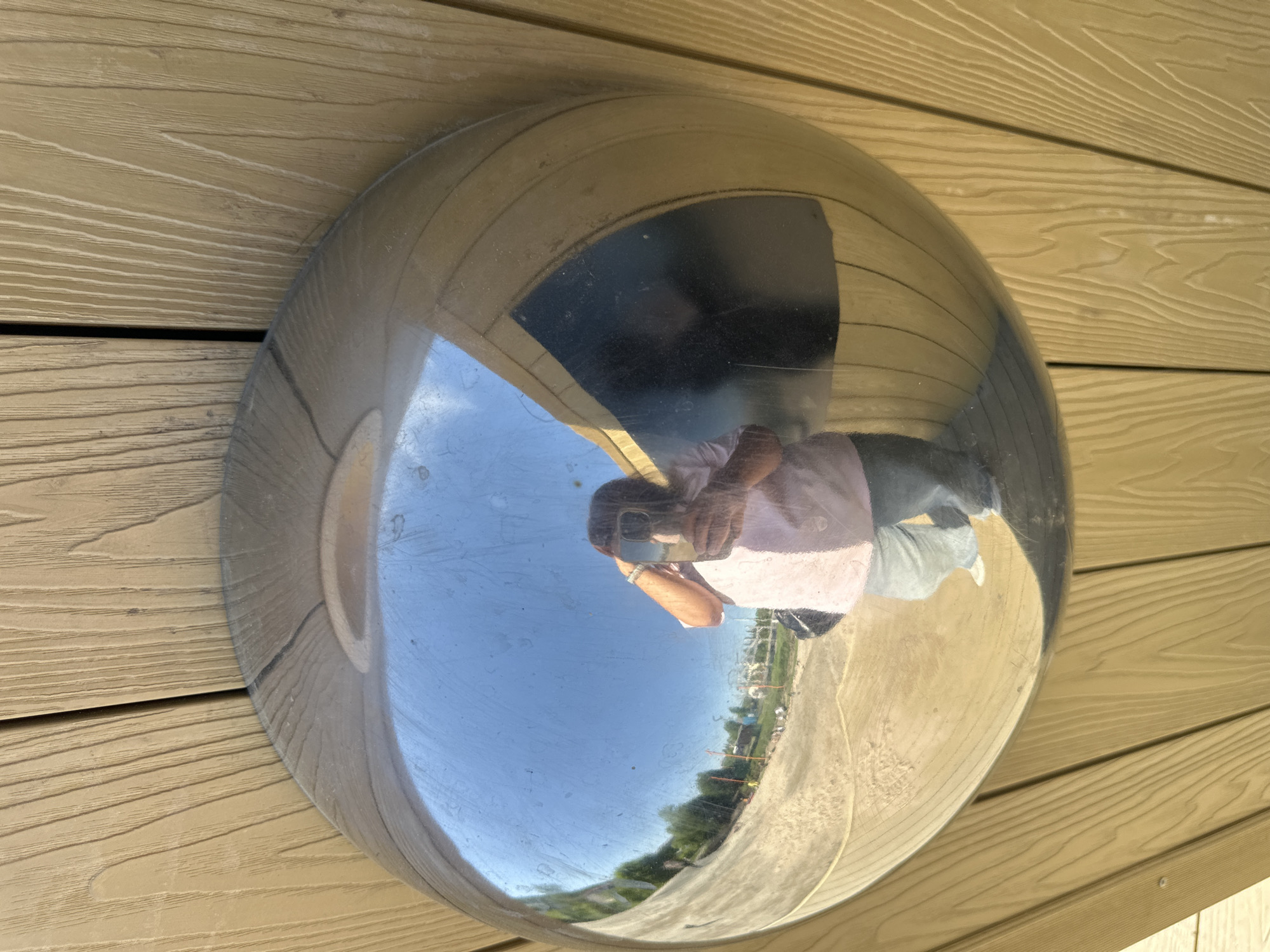 A rounded, metallic convex mirror is mounted on a light brown, horizontally-laid wooden surface. The mirror reflects a distorted scene that includes a person holding a cell phone, likely taking a photograph of themselves. Reflected in the mirror's surface is a blue sky with sparse clouds, buildings, trees, and ground, suggesting an outdoor environment. The city is Unknown, Unknown.