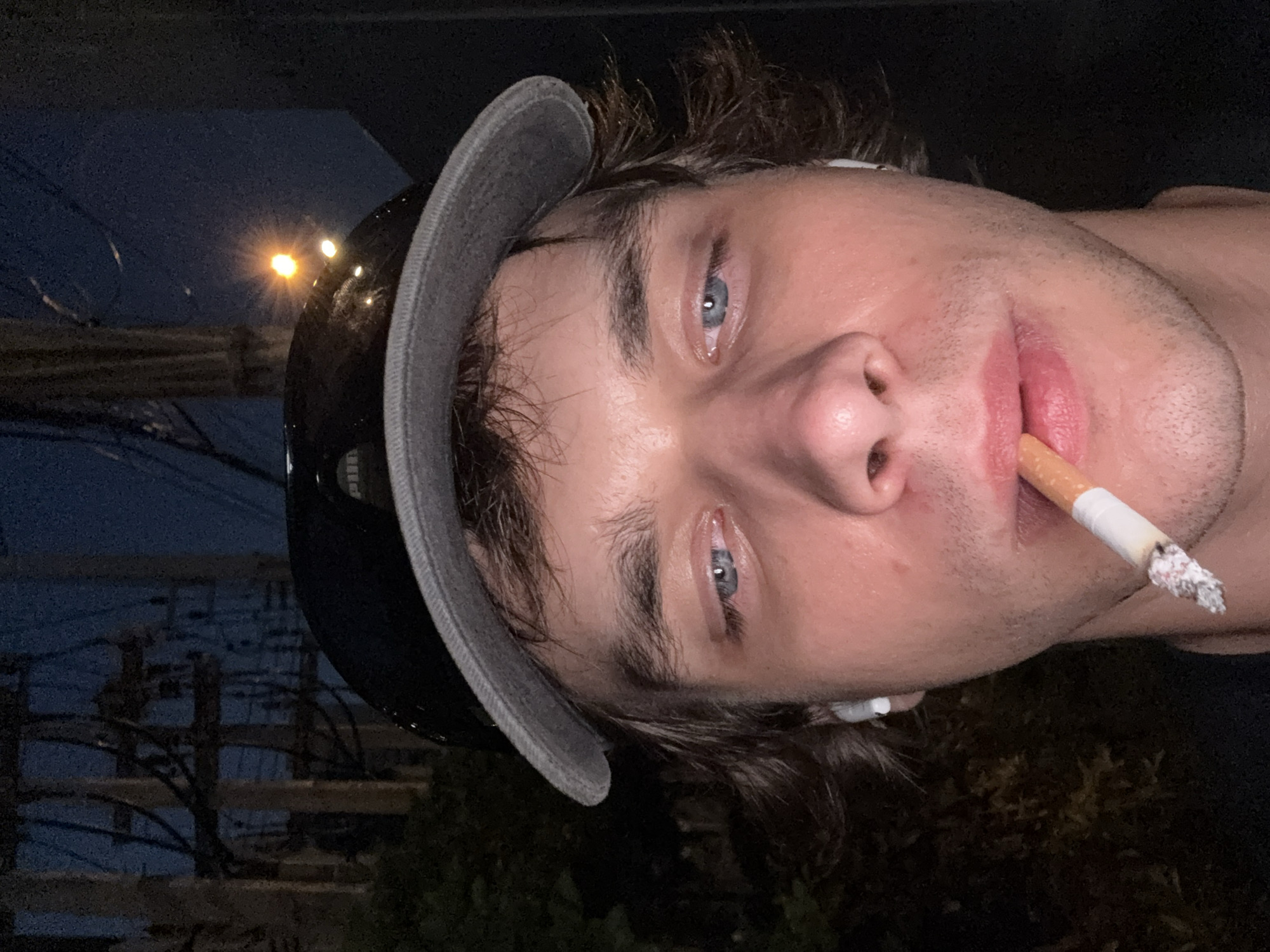 A close-up portrait shows a young man with light skin and blue eyes holding a lit cigarette in his mouth. He wears a black helmet with a gray brim. A wireless earbud is visible on the side of his face. The background suggests an urban environment at night. Power lines are visible along with a distant street light. It is not possible to confirm the location of the scene.