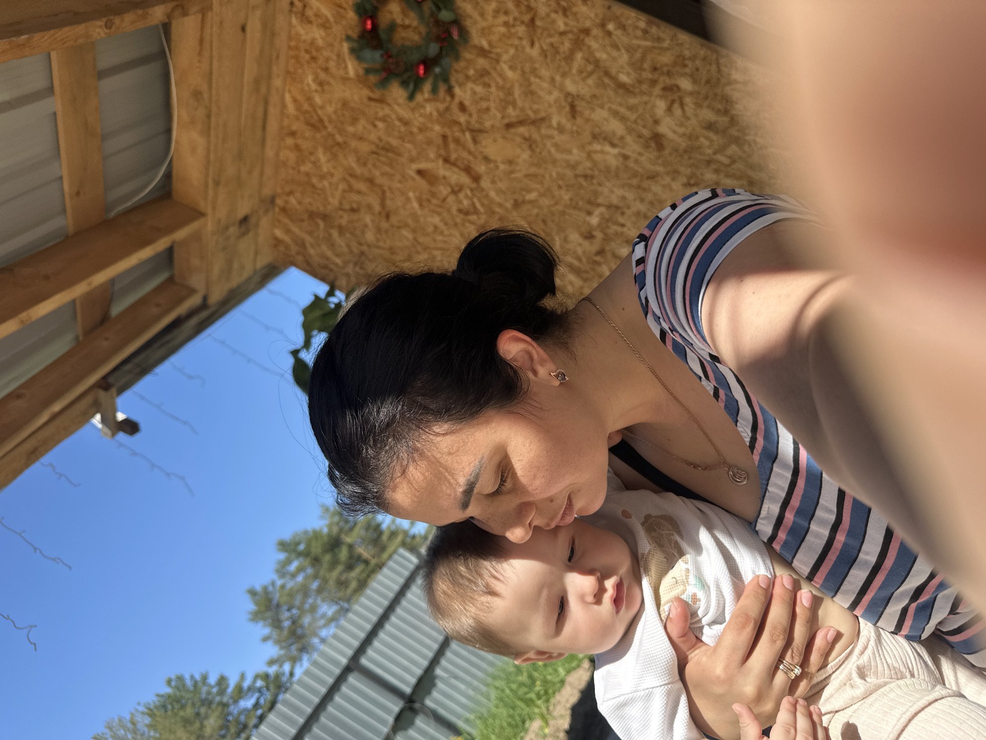 A medium shot captures a woman holding a baby outside on a sunny day. The woman is wearing a striped top and a necklace. Her dark hair is pulled back in a bun. The baby, dressed in white, is held close to her. They are under a wooden structure with a wreath hanging from the roof. The background includes a blue sky and greenery. The location appears to be outdoors, with the wooden structure providing partial shade.