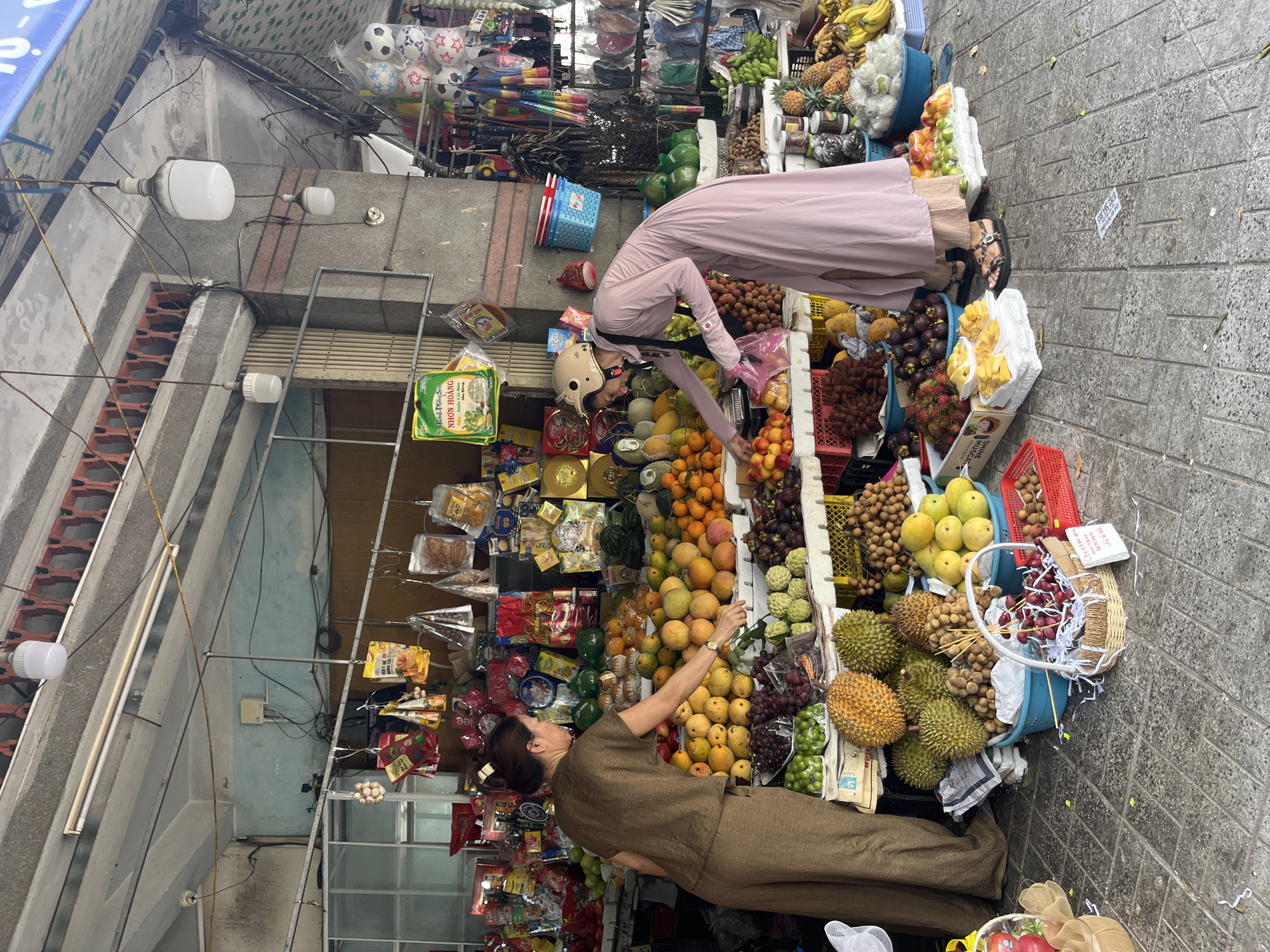 The image depicts an outdoor fruit stand on a tiled sidewalk in Nha Trang Ward, Vietnam. Two women are present, one in a brown tunic and pants, and the other in a long pink dress and a helmet, suggesting they may have arrived by motorcycle. The woman in brown is standing behind the fruit stall, while the woman in pink is inspecting fruit. The stall is laden with a variety of fresh fruits, including durian, longan, mangoes, pineapples, and bananas, displayed in baskets and crates. Packaged goods, such as snacks and dried fruits, are also visible on shelves and hanging from the stall's structure. The scene is characterized by a busy, market-like atmosphere, with overhead lighting and awnings providing shade.