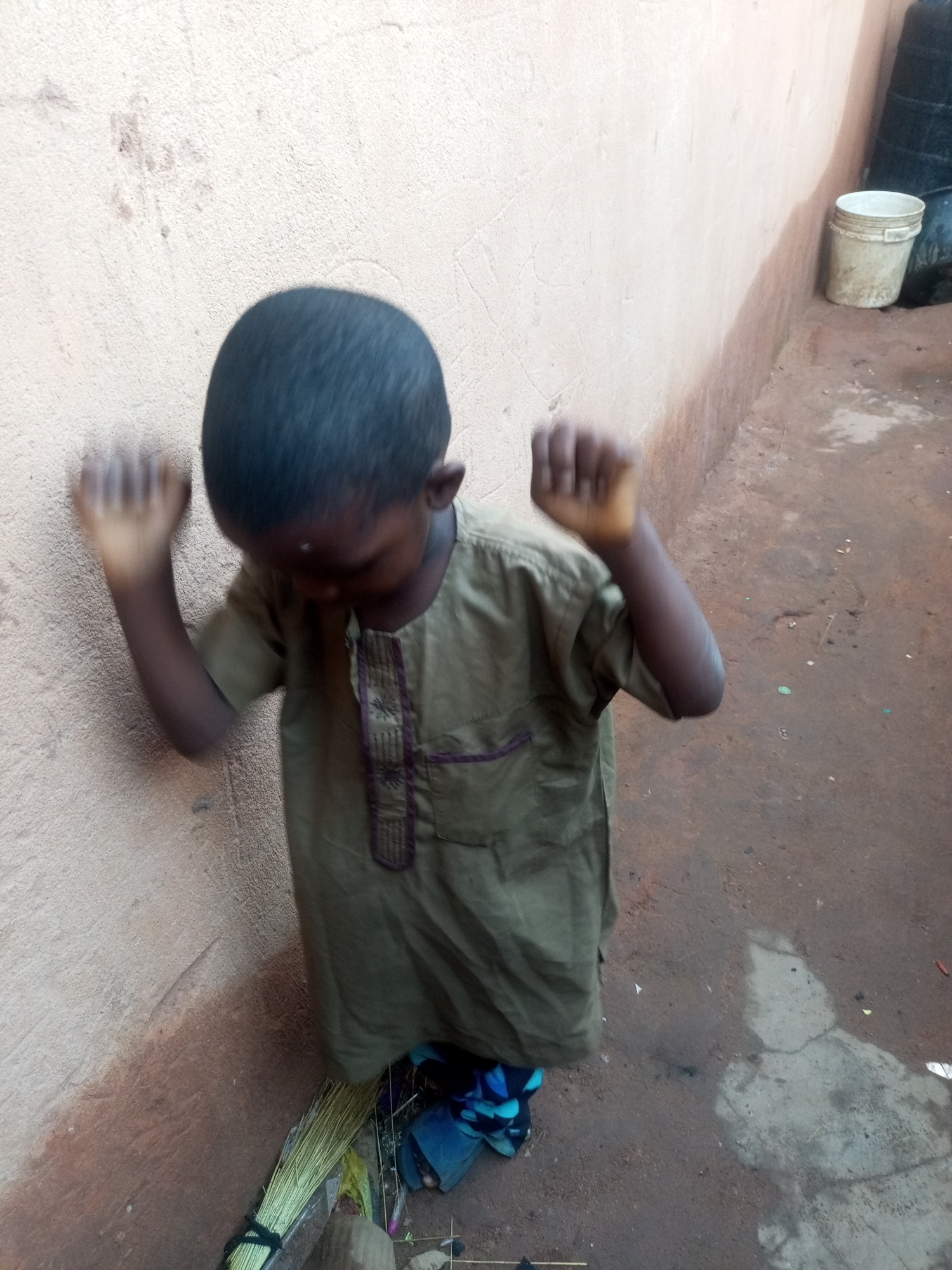 A young boy, appearing to be of African descent, stands against a light brown, textured wall. He is wearing a loose-fitting, olive-green garment with decorative stitching in purple and brown around the neckline. His head is bowed, and his hands are raised and clenched into fists, positioned near the wall as if he is leaning on it or being reprimanded. The boy's hair is short and dark.
The environment appears to be an outdoor, likely urban or semi-urban setting. The ground is a reddish-brown dirt, uneven and cracked in places. To the right of the boy, a weathered white plastic bucket stands near the wall. Further back, a dark, possibly black, cylindrical object is visible, partially obscured. A bundle of dry grass, tied with black string, lies on the ground near the boy's feet, along with some other debris including a blue cloth. The overall impression is of a humble and somewhat neglected environment, possibly in the city of Tempure, Nigeria.