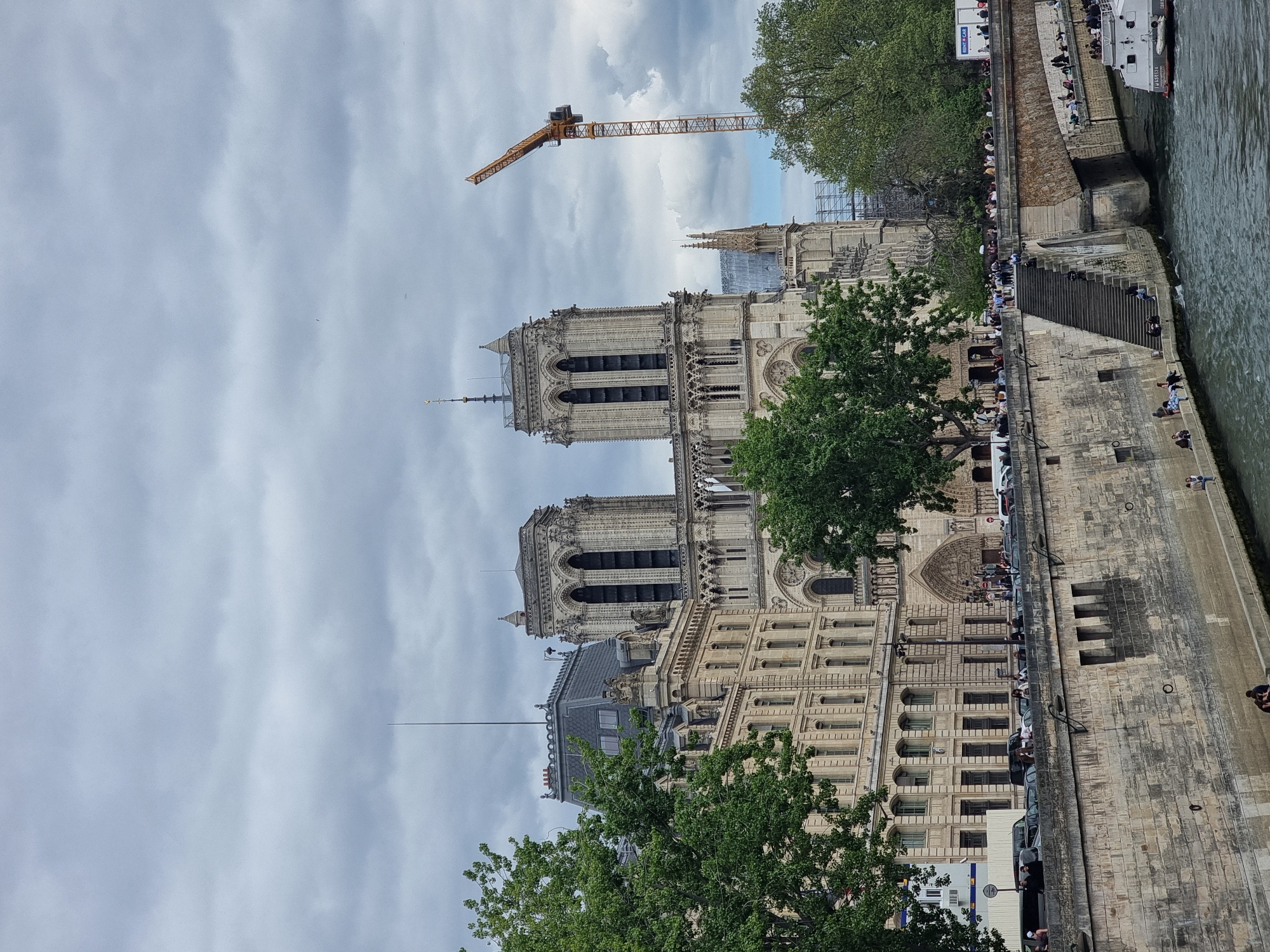 The image shows Notre Dame Cathedral in Paris, France. The cathedral's iconic towers are visible, along with a construction crane in the background. People are gathered along the riverbank, and a tour boat is present on the water. The sky is overcast.