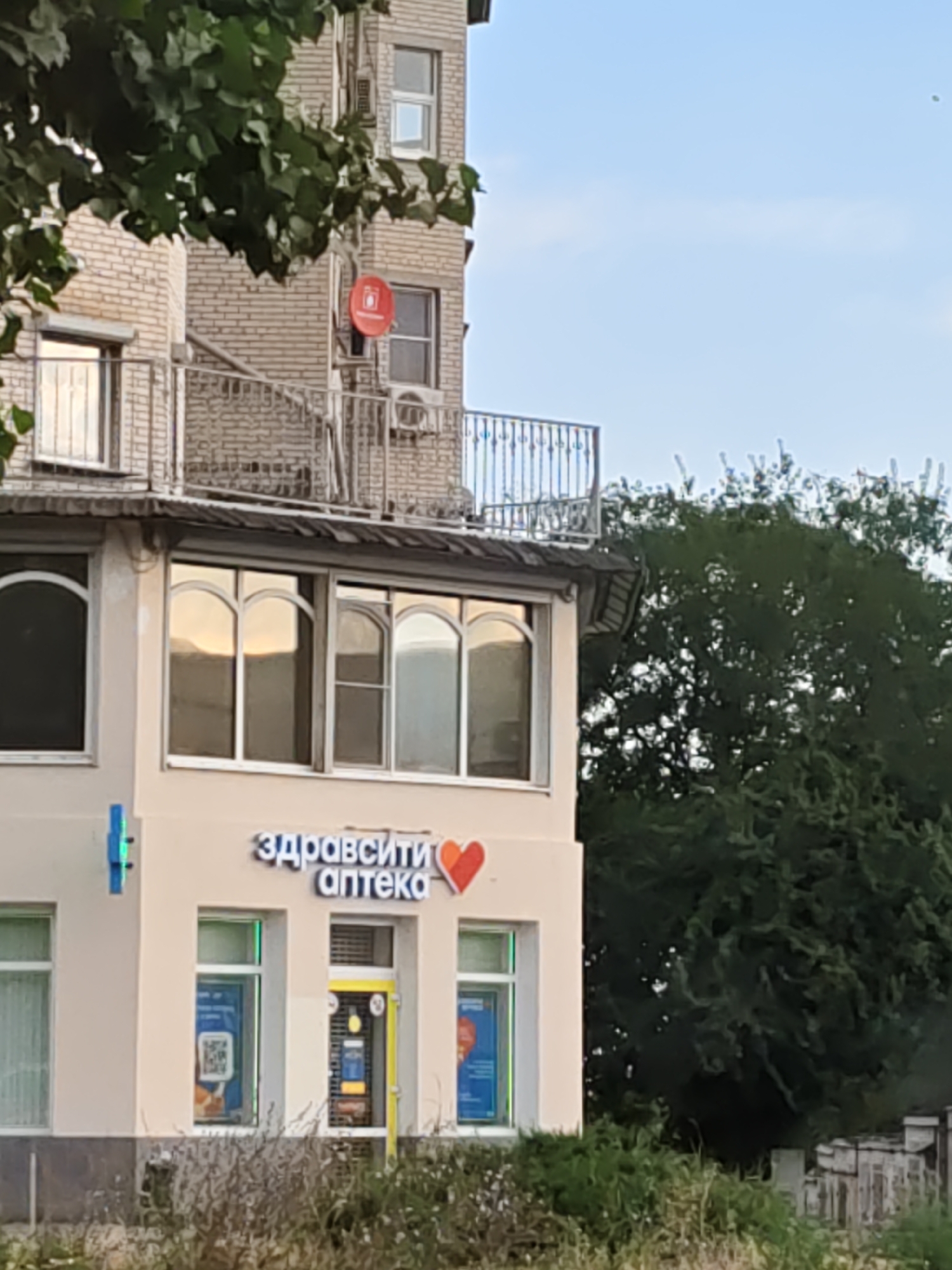 The image shows the facade of a building in Novocherkassk, Russia, housing an "Apteka" (pharmacy). The pharmacy's sign, in Cyrillic, reads "Здравствуйте аптека" which translates to "Hello pharmacy." A red heart symbol is integrated into the lettering of the sign. The pharmacy has large display windows, one of which features a QR code. A blue cross-shaped pharmacy sign is visible to the left of the entrance. The building has a light-colored facade and arched windows on the upper floor. A large, leafy tree dominates the right side of the image, with dense green foliage. The sky above is a clear blue with some wispy clouds. The lower portion of the image shows dry grass and some small shrubs. In the background, a multi-story brick building is partially visible, with balconies and what appears to be a satellite dish.