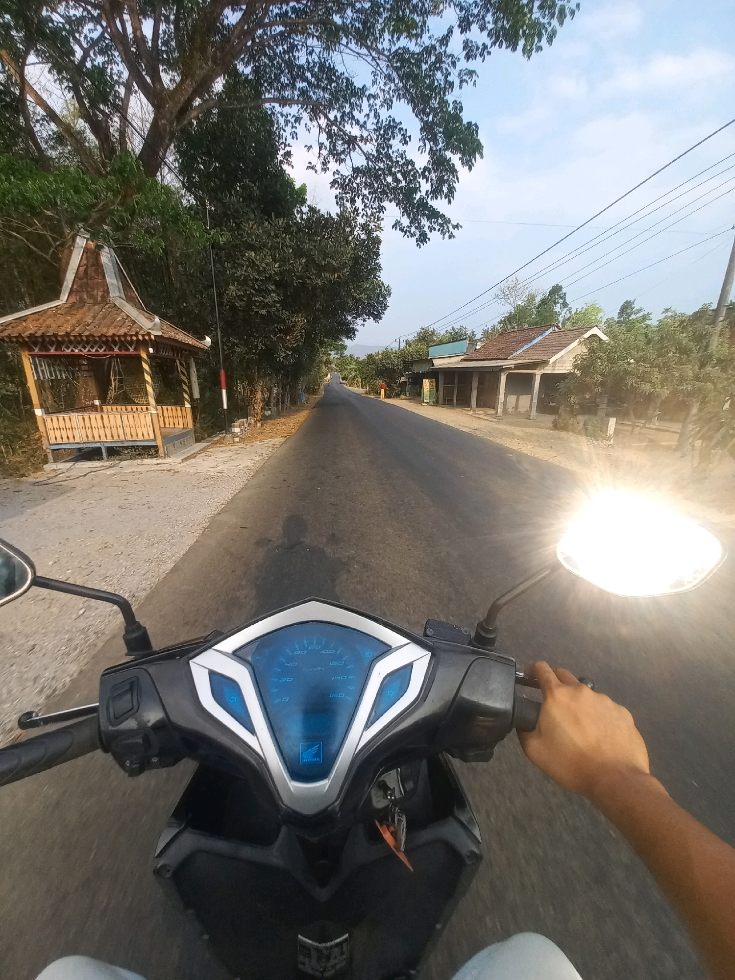 A first-person perspective shot from a motorcycle in Ngarjosari, Indonesia, shows a road bordered by trees and residential structures. The road ahead is asphalt and curves gently to the right. On the left side of the road, there's a small, traditional gazebo with a tiled roof and wooden railings. Further down the road, on the right, are several single-story houses with tiled roofs and open porches. The sky is partly cloudy and bright, with the sun glinting intensely off the motorcycle's right-side mirror, creating a lens flare. The motorcycle's dashboard with a blue digital display is visible in the foreground, and a person's hand is gripping the handlebar. The overall scene depicts a rural or semi-rural setting typical of Indonesian villages.