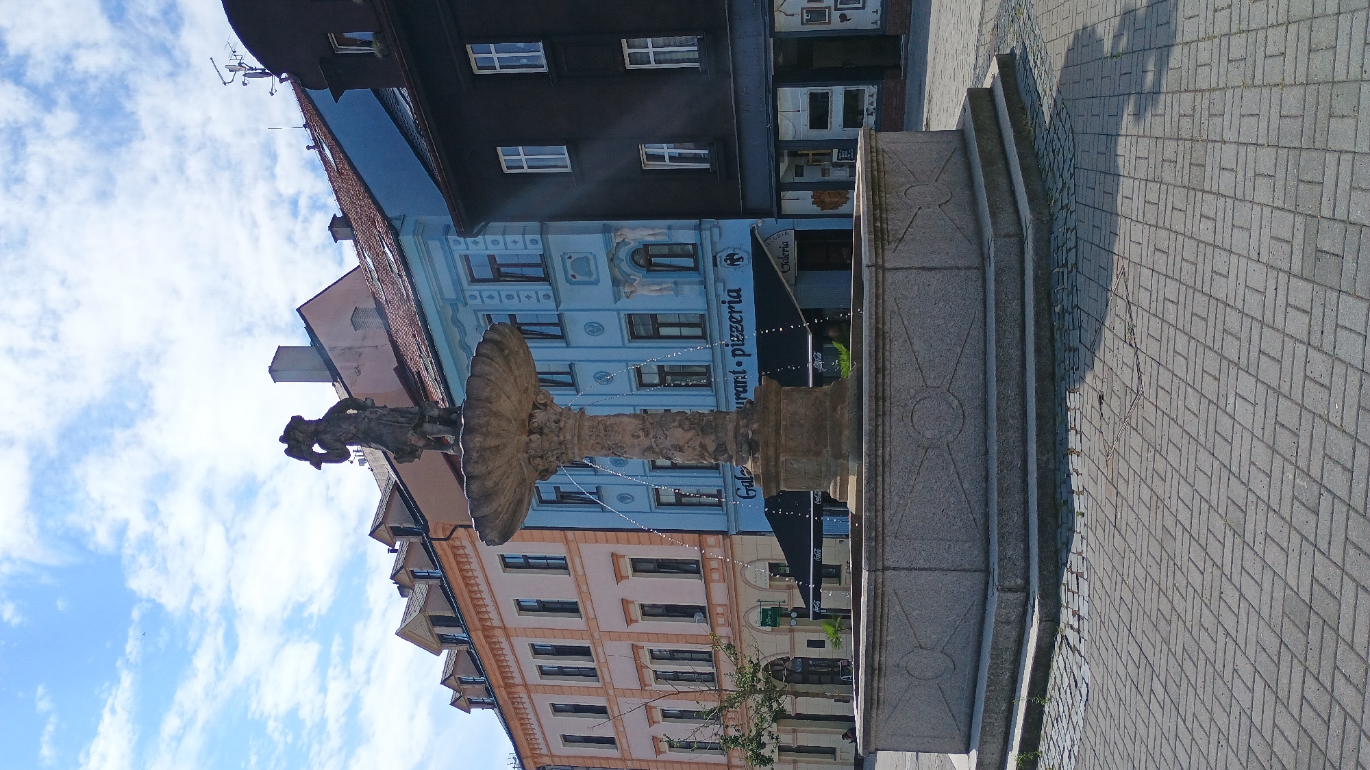 In the city of Sokolov, Czechia, a stone fountain stands in a paved square. The fountain features a multi-tiered basin with water cascading from the top. A weathered statue of a person holding a basket or headdress adorns the summit. The square is surrounded by colorful buildings, with a pale orange building on the left, a bright blue building in the center, and a dark building on the right. The blue building has signage for "Galeria" and "Restaurant-Pizzeria." The ground is paved with interlocking rectangular stones. The sky above is blue with scattered white clouds.