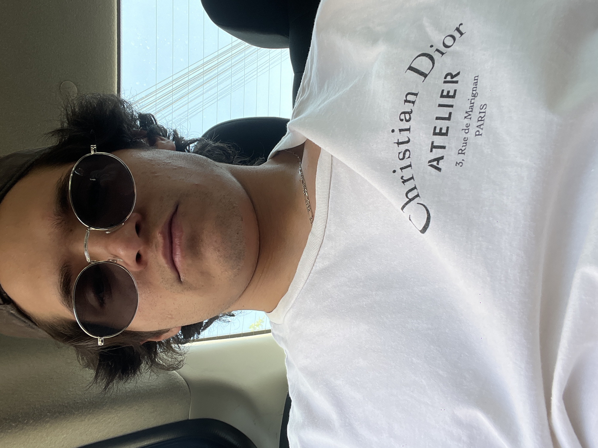 An eye-level, close-up portrait captures a young man seated in what appears to be the interior of a vehicle during the daytime. He looks directly at the viewer with a neutral expression. He has dark, curly hair and visible stubble on his chin and jawline. He wears dark, round-lensed sunglasses with silver frames that sit low on his nose. A silver Figaro chain necklace is visible around his neck. He is dressed in a plain white t-shirt, on which bold black text reads: "Christian Dior ATELIER 3, Rue de Marignan PARIS". The background, seen through a vehicle window, reveals a bright, slightly overcast sky with several thin, parallel lines running horizontally across it, possibly power lines or reflections. The immediate surroundings within the vehicle are dark and simple, with a dark headrest visible. The scene suggests a casual moment in Mexico City, Mexico.