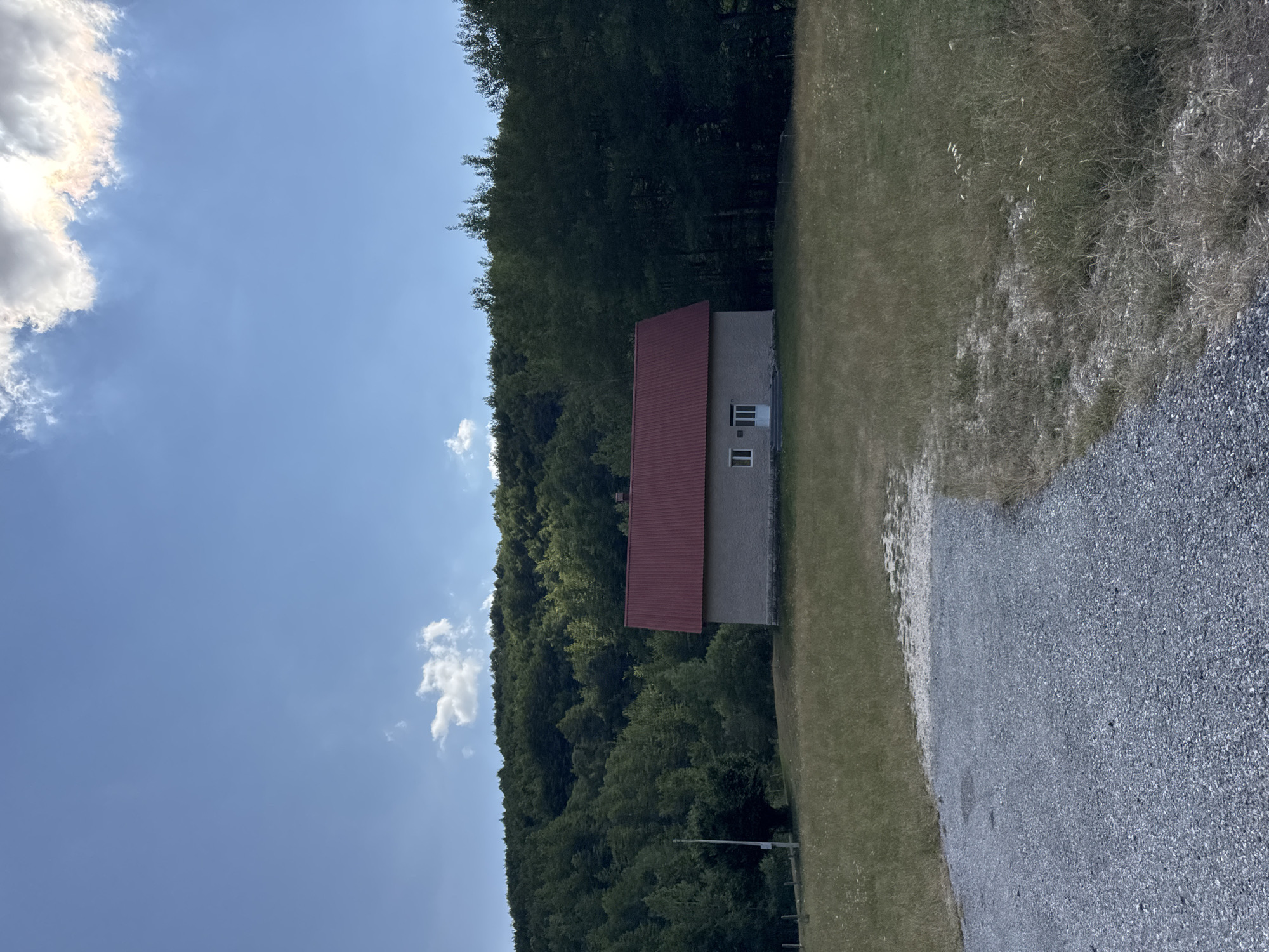 The image shows a small, single-story building with a red-tiled roof, situated in a grassy field. The building has light-colored stone walls and two windows on the facade. A gravel path leads from the foreground towards the building. Behind the building, a dense forest covers a hillside, with a clear blue sky above, featuring scattered white clouds. The scene appears to be in a rural or mountainous area, possibly in Donja Bukovica, Montenegro, given the context. There are no people or animals visible in the image. The environment is natural, with vegetation and a clear sky.