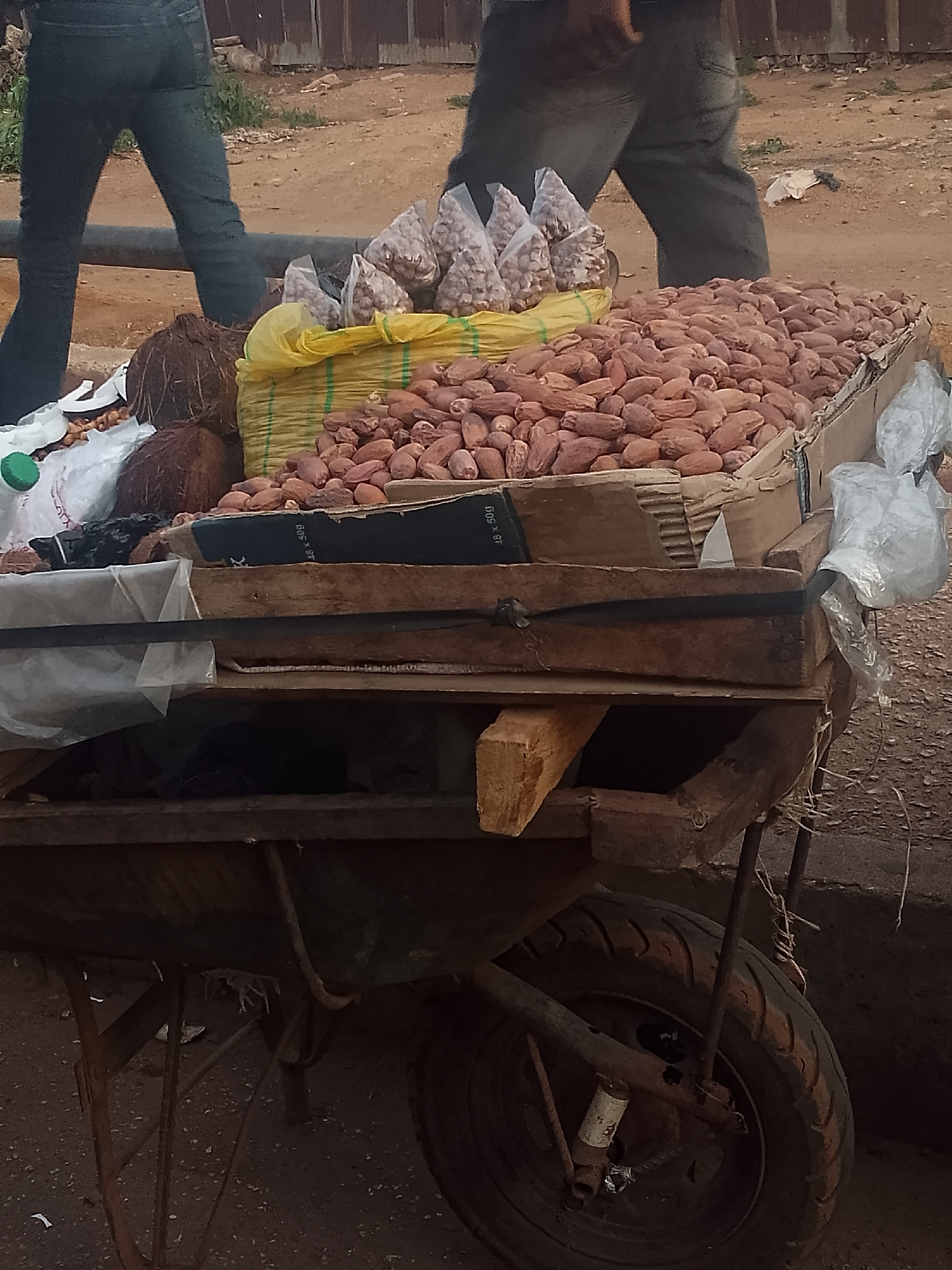 A street vendor's wheelbarrow in Bukuru, Nigeria is heavily loaded with goods for sale. The wheelbarrow itself appears aged and weathered, with a single worn tire and rusty metal supports. The cart's wooden frame is layered with cardboard, forming a makeshift container.
The primary merchandise is a large quantity of dates, piled high within the cardboard-lined container. Beside the dates are two coconuts and several small plastic bags containing what appear to be nuts or seeds. These smaller items are held in a yellow, green and white striped bag. A clear water bottle and some white plastic bags rest on the opposite side of the container.
Two figures are partially visible in the background. One is wearing blue jeans, and the other is wearing gray pants. The ground is dirt or gravel. A metal fence can be seen in the background. The overall setting suggests a bustling, open-air marketplace or street vending area.