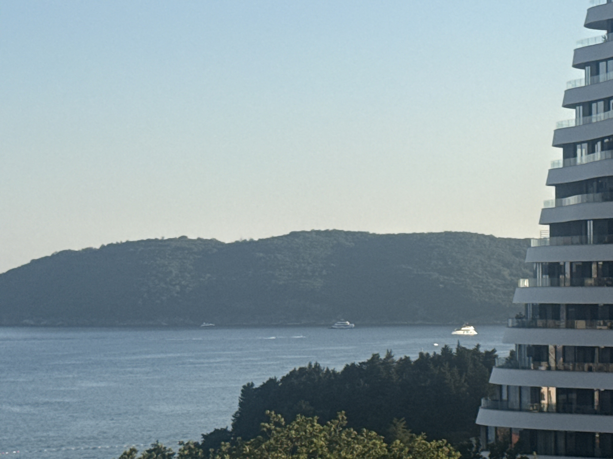 The image shows a view of the Adriatic Sea from Boreti, Montenegro. In the foreground, a modern, multi-story building with a curved facade and numerous balconies is visible on the right side. Beyond the building, the calm, blue sea stretches to the horizon. A tree-covered hill, likely an island or a peninsula, forms the backdrop. Several boats, including a white yacht, are dotted on the water. The sky is clear and a pale blue, suggesting a sunny day.