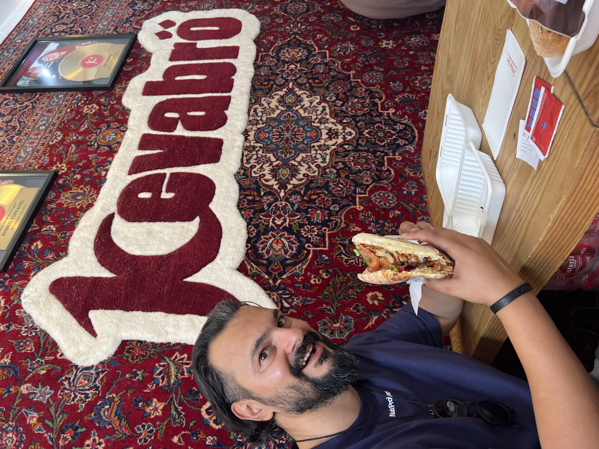 A man with dark hair, a beard, and a blue t-shirt smiles at the camera while holding a large sandwich filled with meat, vegetables, and sauce. He is seated at a wooden table in front of a wall decorated with a large Persian rug and a prominent sign that reads "Kevabro" in white and red lettering. Two white takeout containers and a napkin are on the table, along with some packets of condiments. The man's left hand is visible, wearing a black bracelet. The background wall also features two framed awards or pictures. The overall scene suggests a casual dining experience at a restaurant in Barcelona, Spain.